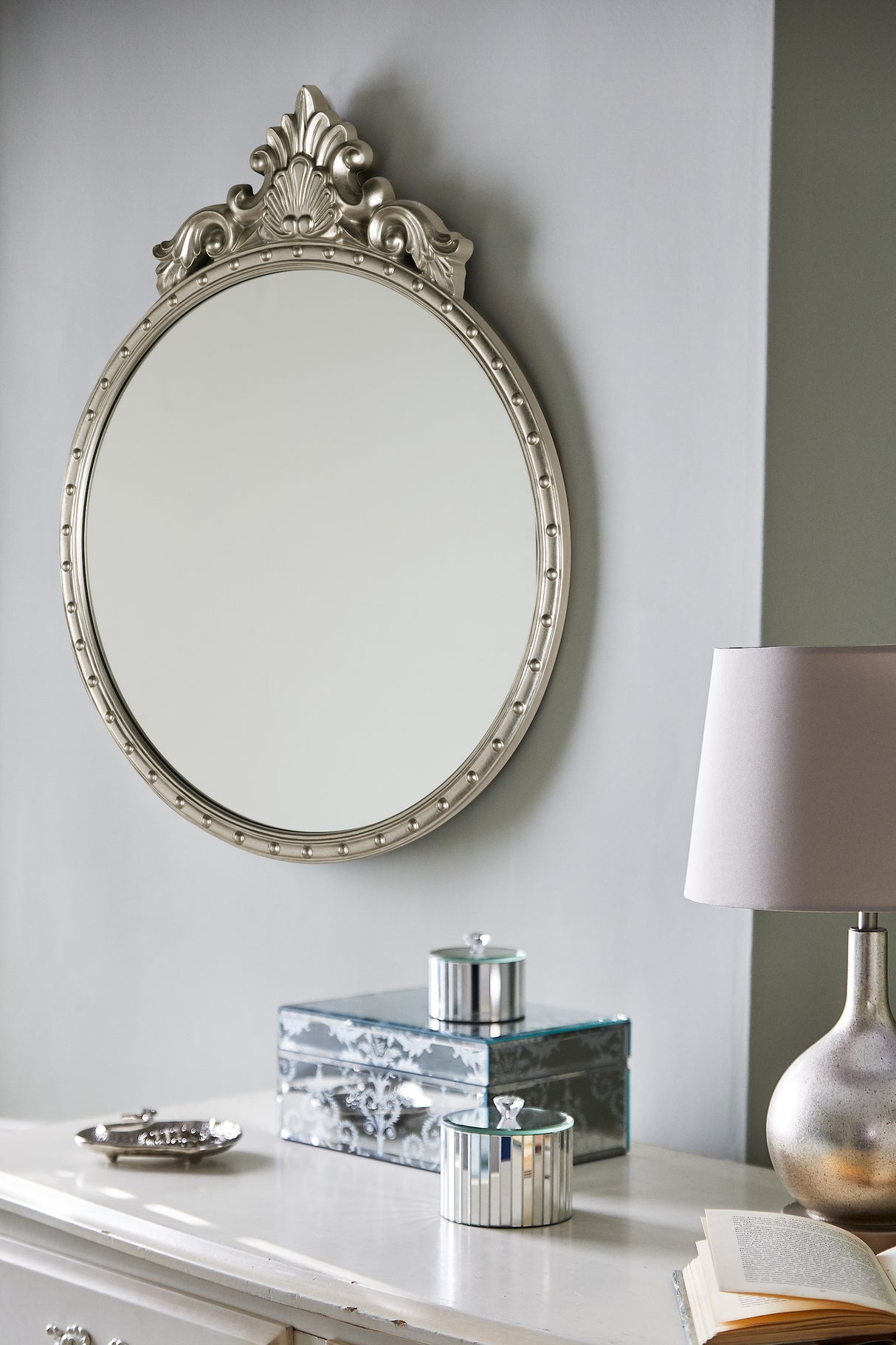 Decorative oval mirror with ornate frame above white dresser with silver boxes, lamp, and open book
