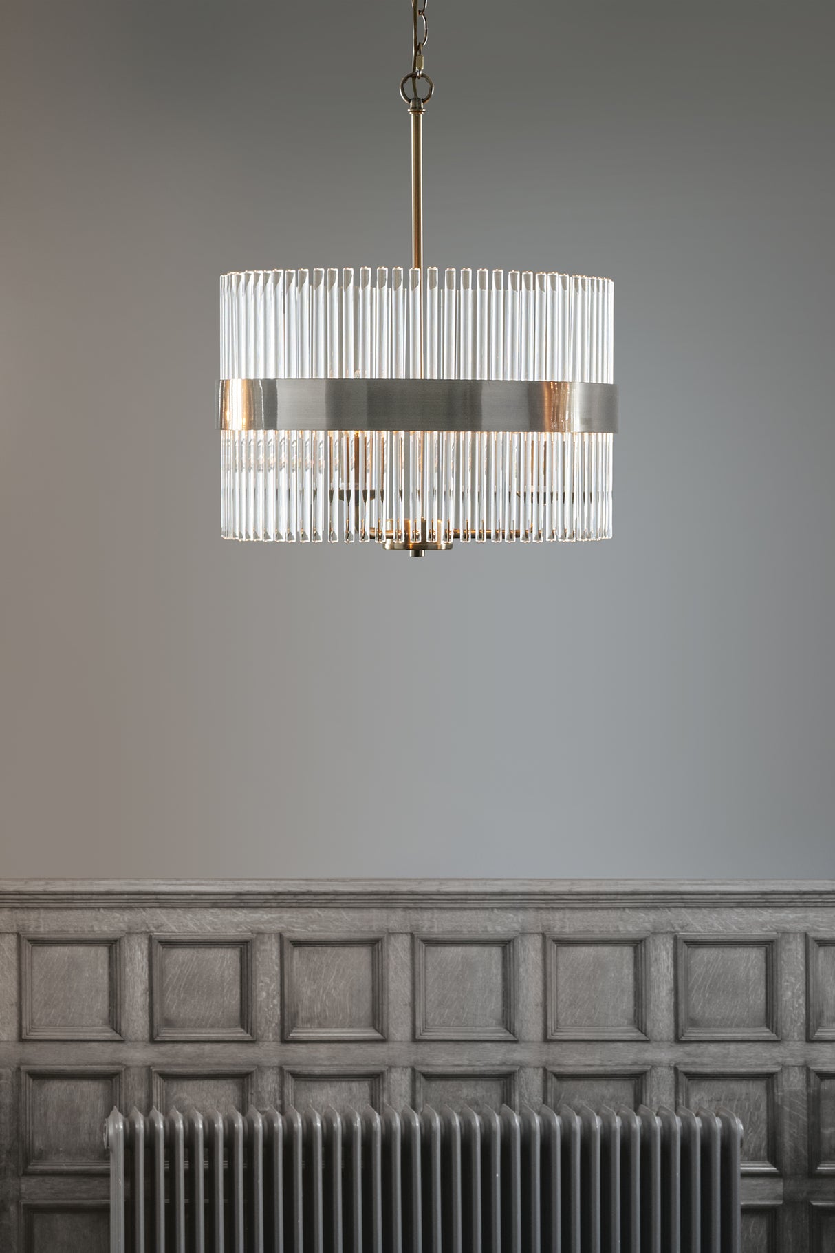 Modern chandelier with glass rods and brass band hanging over gray paneled wall and radiator