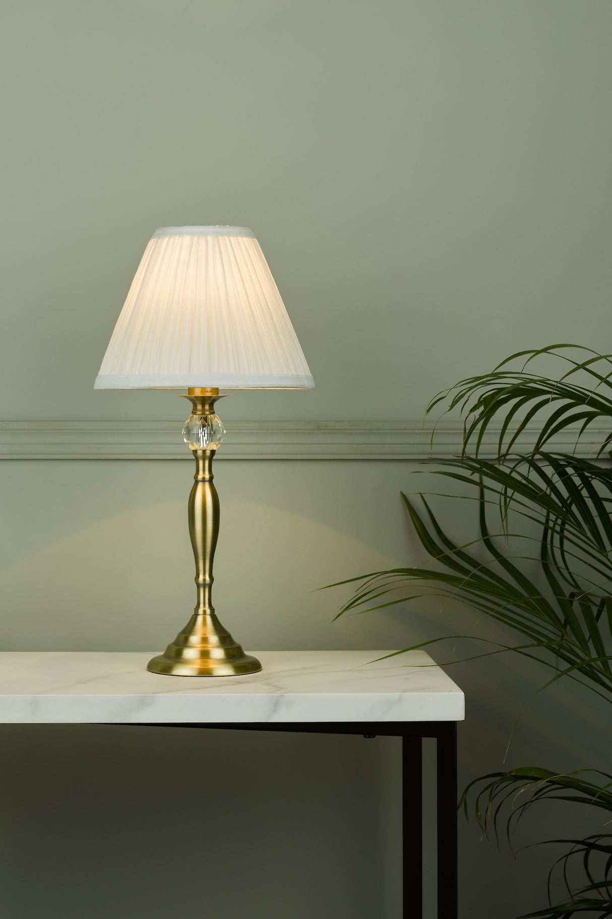 Elegant brass table lamp with white pleated shade on marble-top table beside green plant