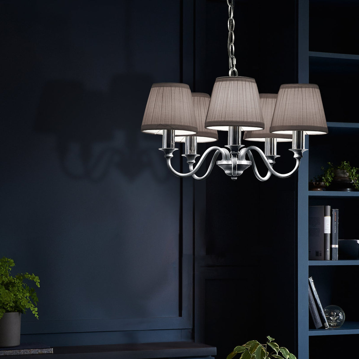 Modern silver chandelier with five white fabric lampshades hanging in a dark blue room with shelves and plants