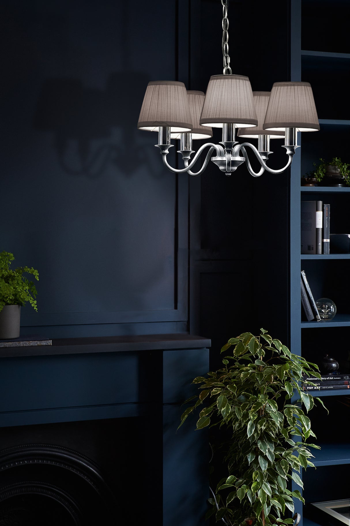 Silver chandelier with fabric lampshades illuminating dark blue interior with green plants and bookshelf