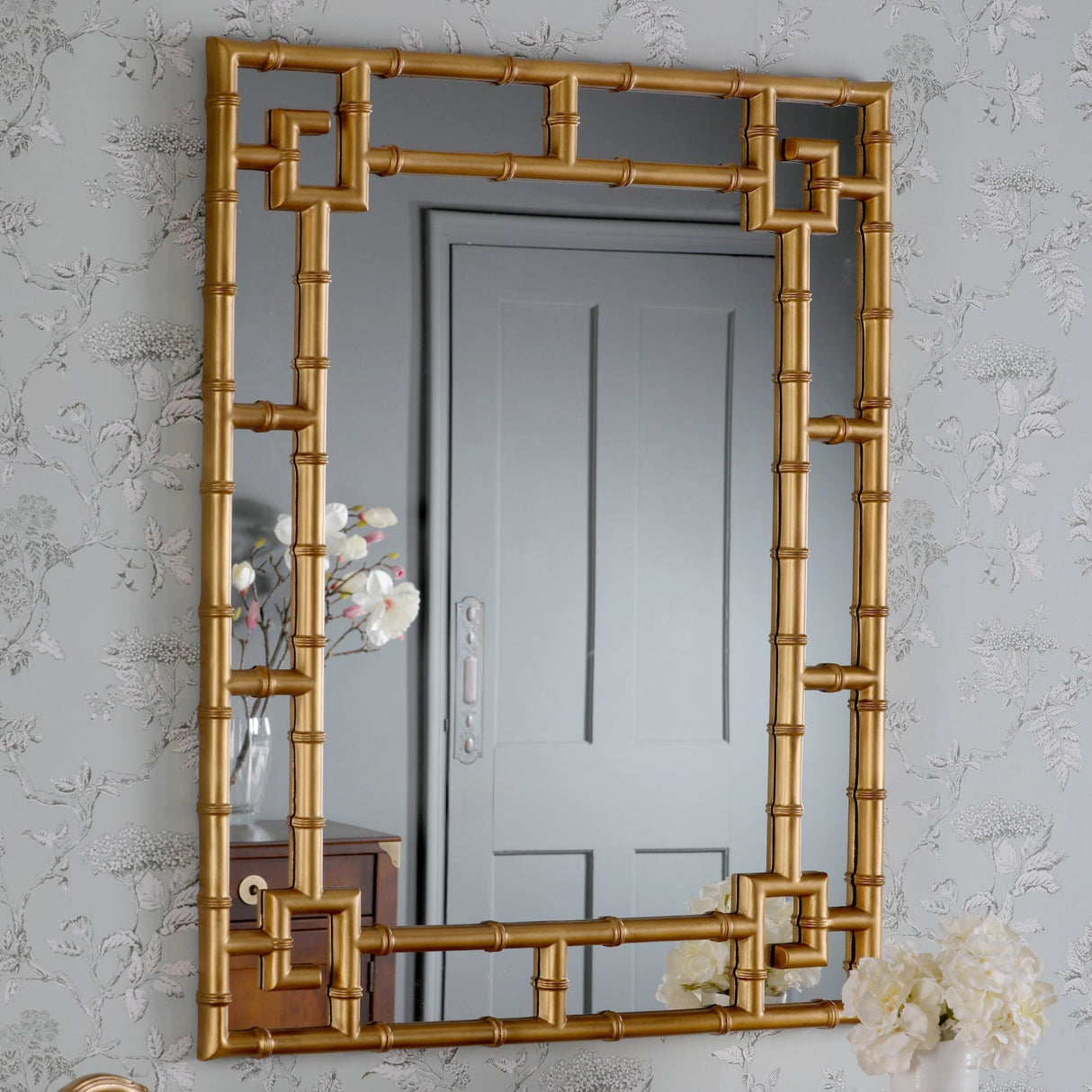 Gold bamboo-style framed mirror reflecting grey door with floral wallpaper in elegant room