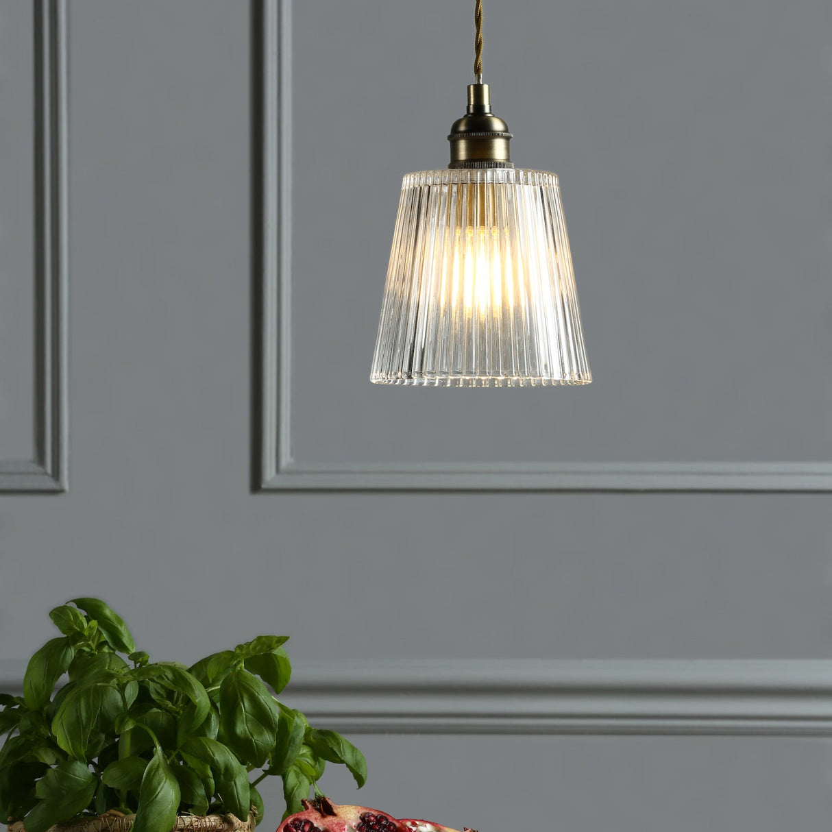 Modern glass pendant light hanging with brass details above a green basil plant against gray paneled wall