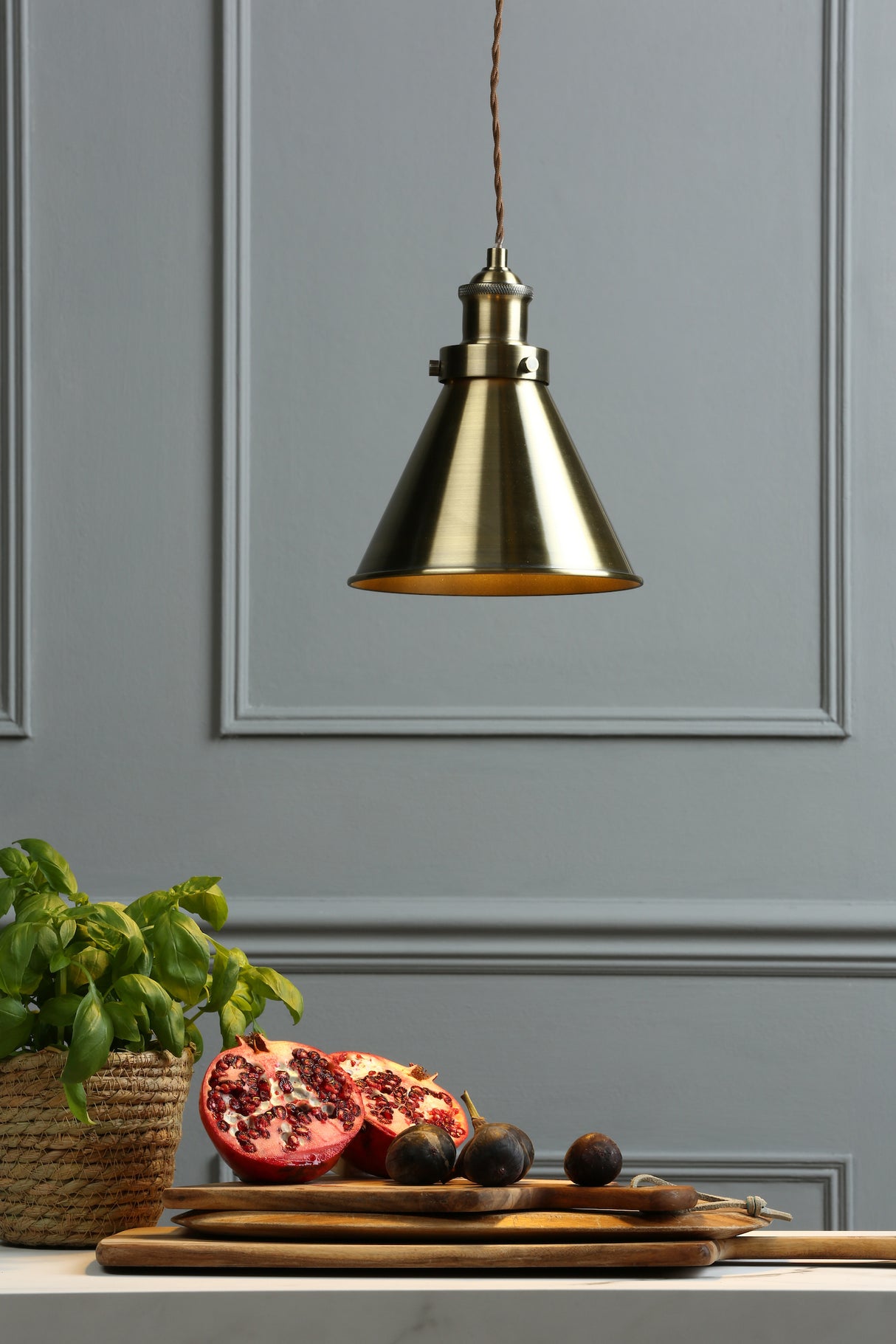 Brass pendant light above wooden cutting boards with pomegranate and figs on gray paneled wall background