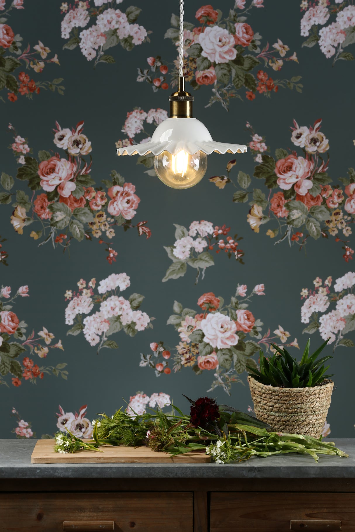 Vintage white pendant light with ruffled edge above flowers on wooden board and potted plant on countertop