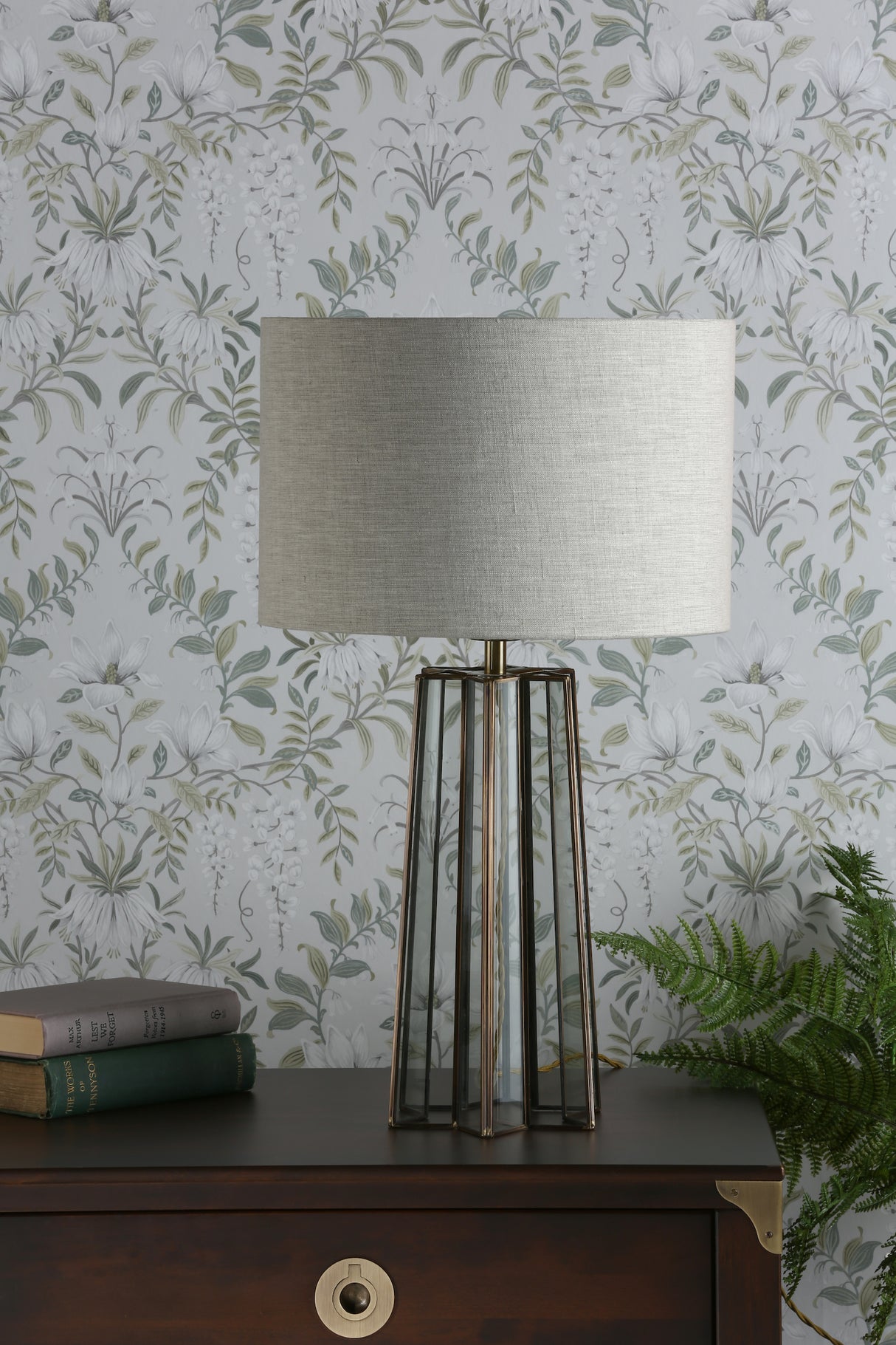 Modern table lamp with beige shade and geometric glass base on wooden dresser with floral wallpaper