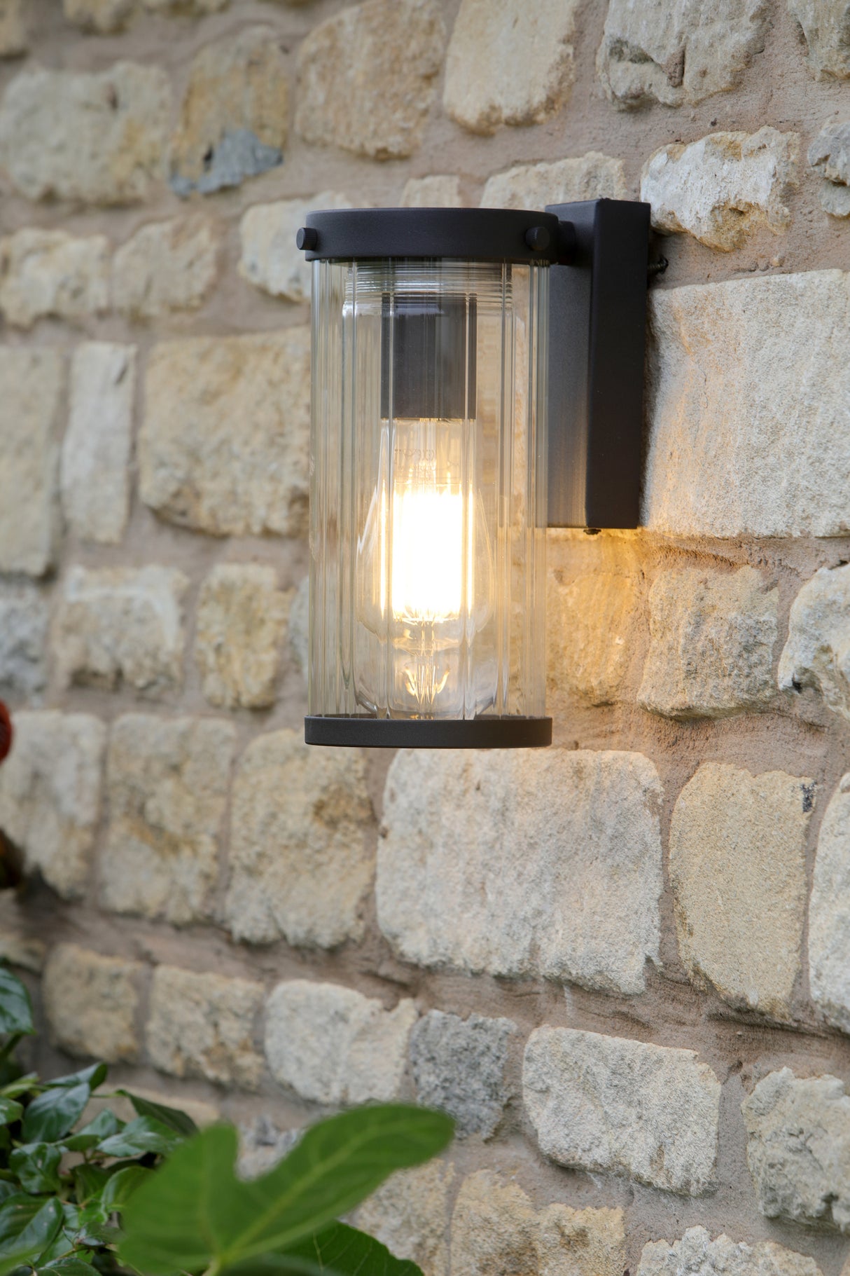 Black outdoor wall lantern with clear ribbed glass mounted on stone brick wall illuminated at dusk