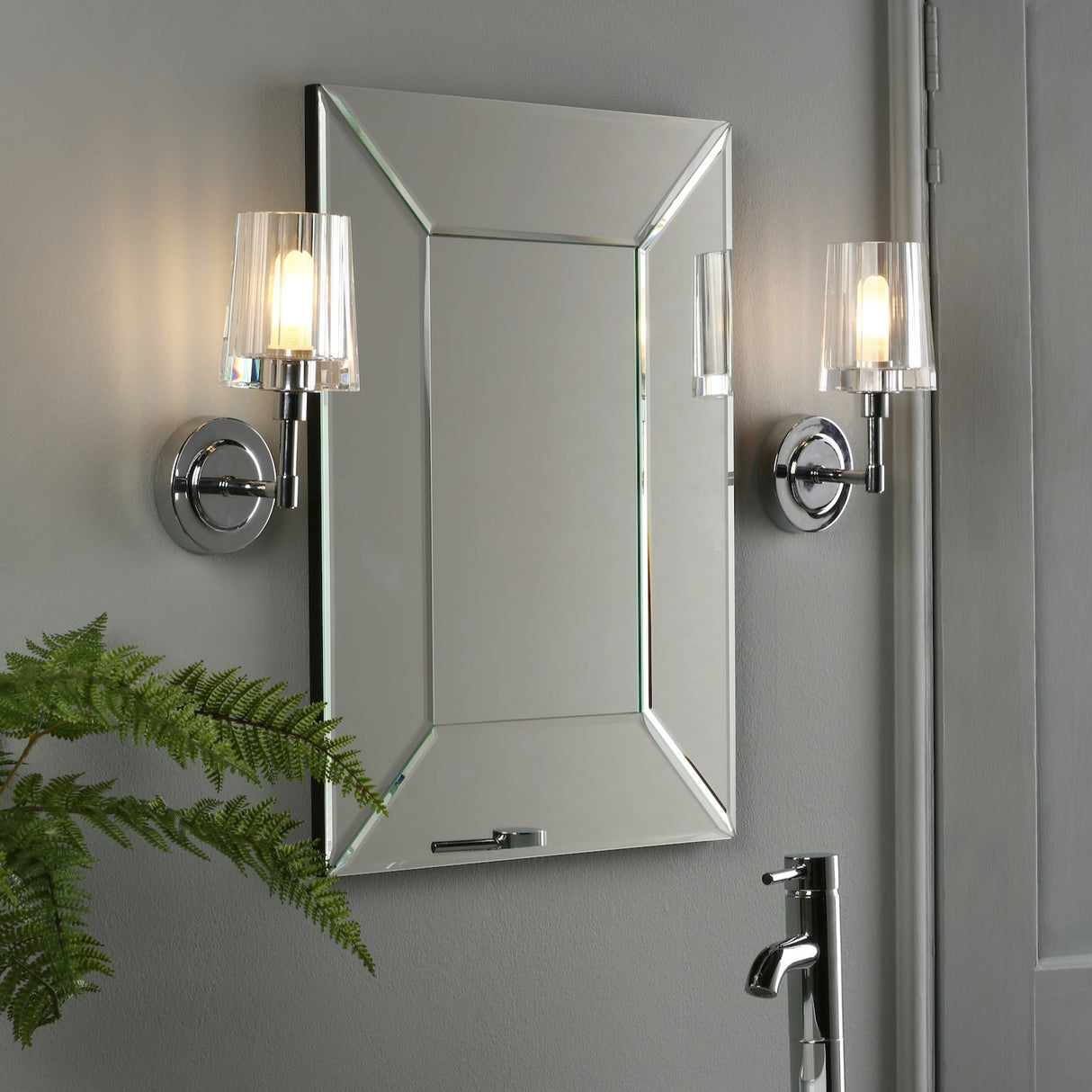 Modern bathroom with beveled mirror, chrome faucet, wall sconces with glass shades, and green fern