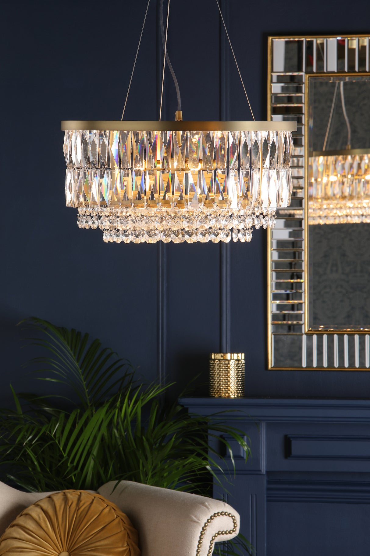 Elegant crystal chandelier with gold accents hanging in navy blue living room with beige sofa and round cushion