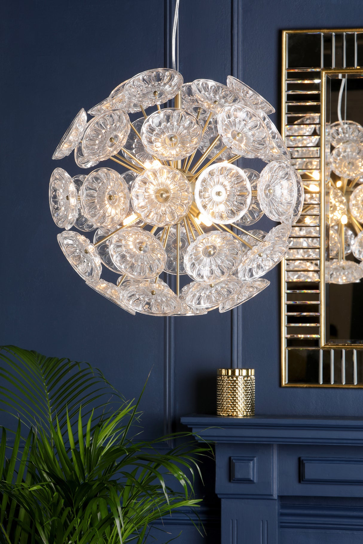 Modern spherical chandelier with clear glass discs against a dark blue wall near a gold mirror and green plant
