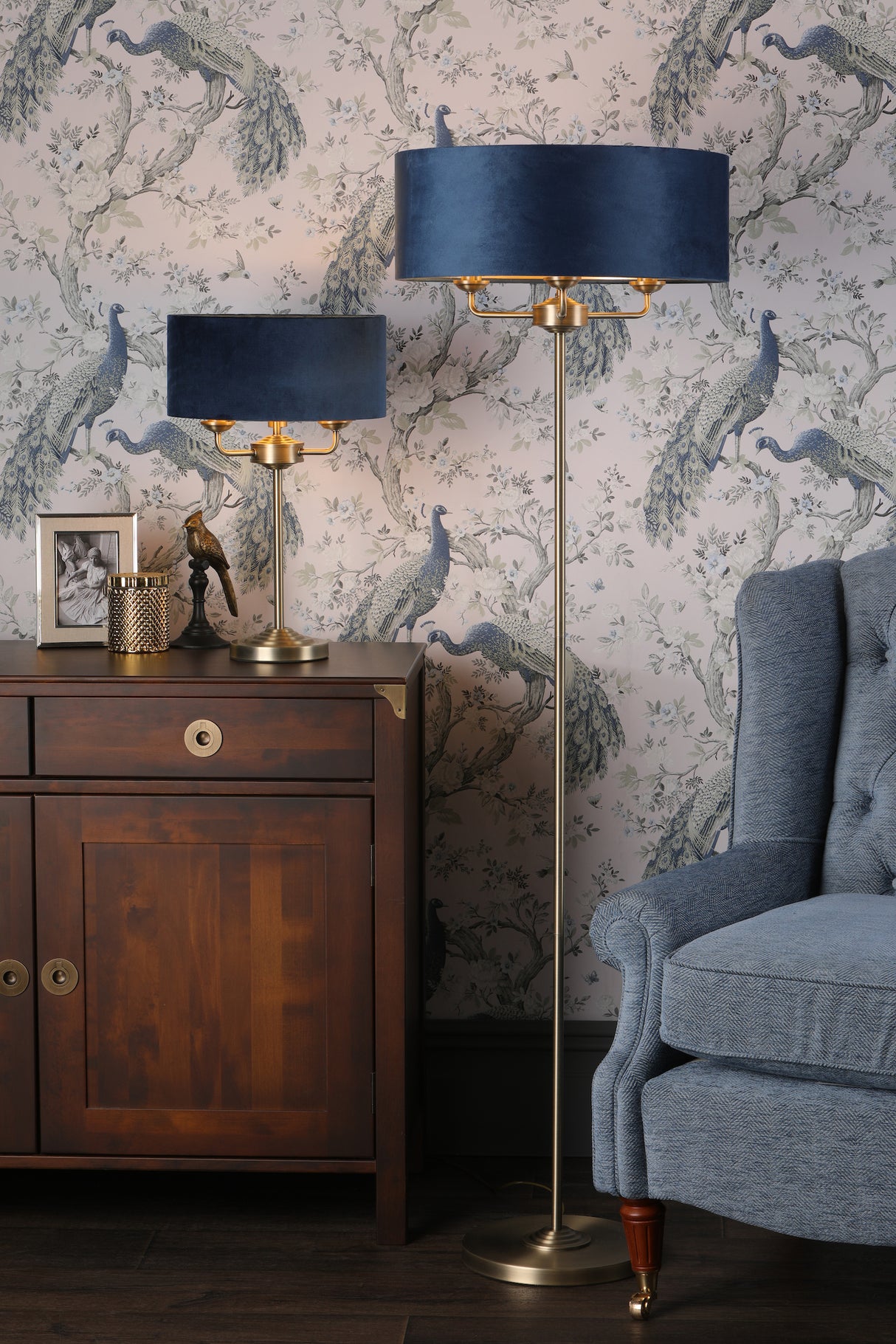 Cozy living room corner with blue velvet lampshades on brass floor and table lamps, dark wood cabinet, blue upholstered armchair, and peacock wallpaper
