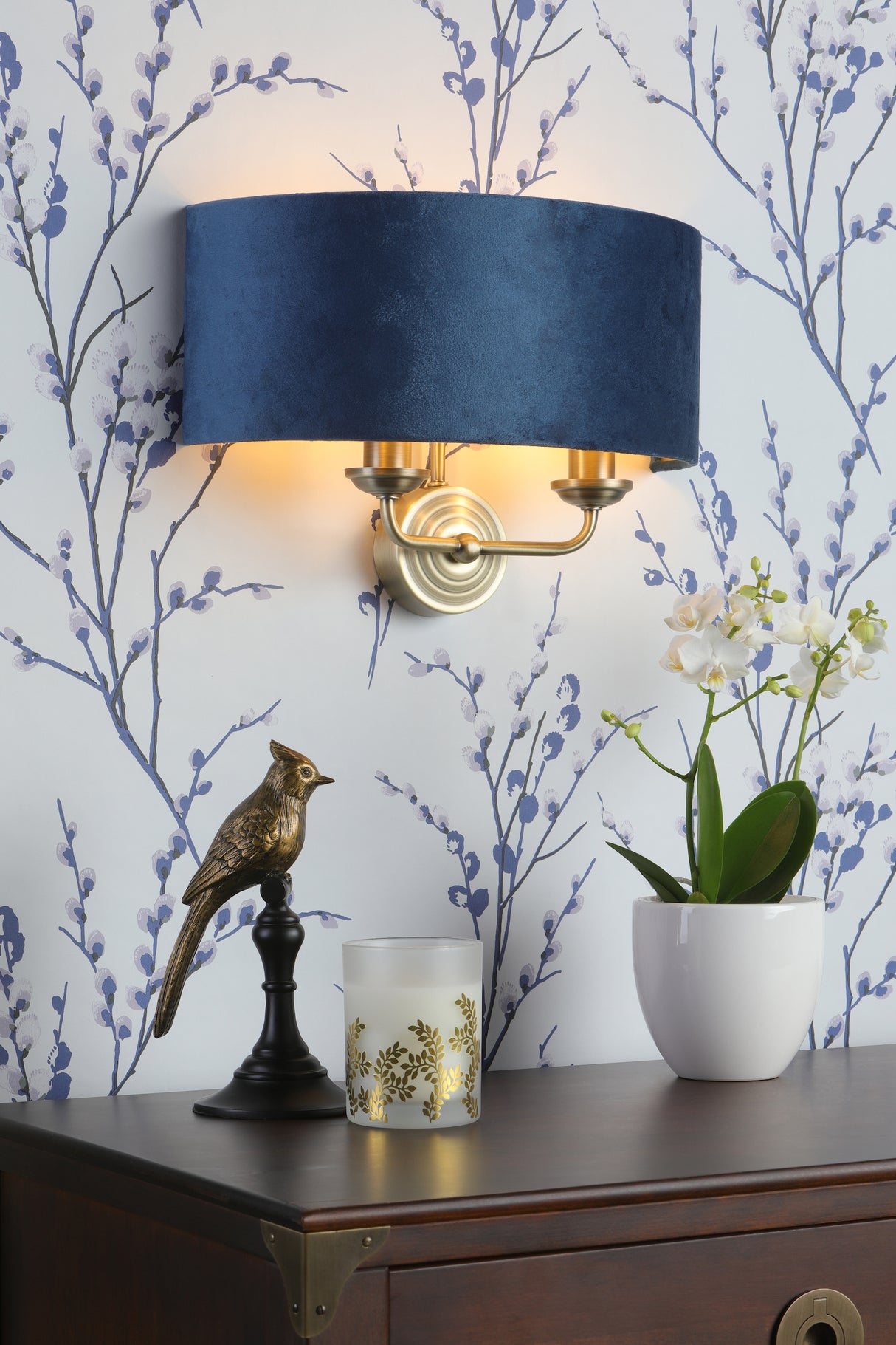 Elegant navy blue wall sconce with gold accents above wooden dresser with bird statue, candle, and white orchid