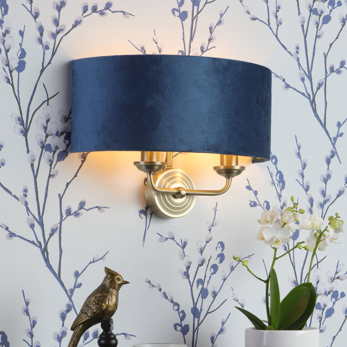 Elegant navy blue wall sconce with gold accents above wooden dresser with bird statue, candle, and white orchid