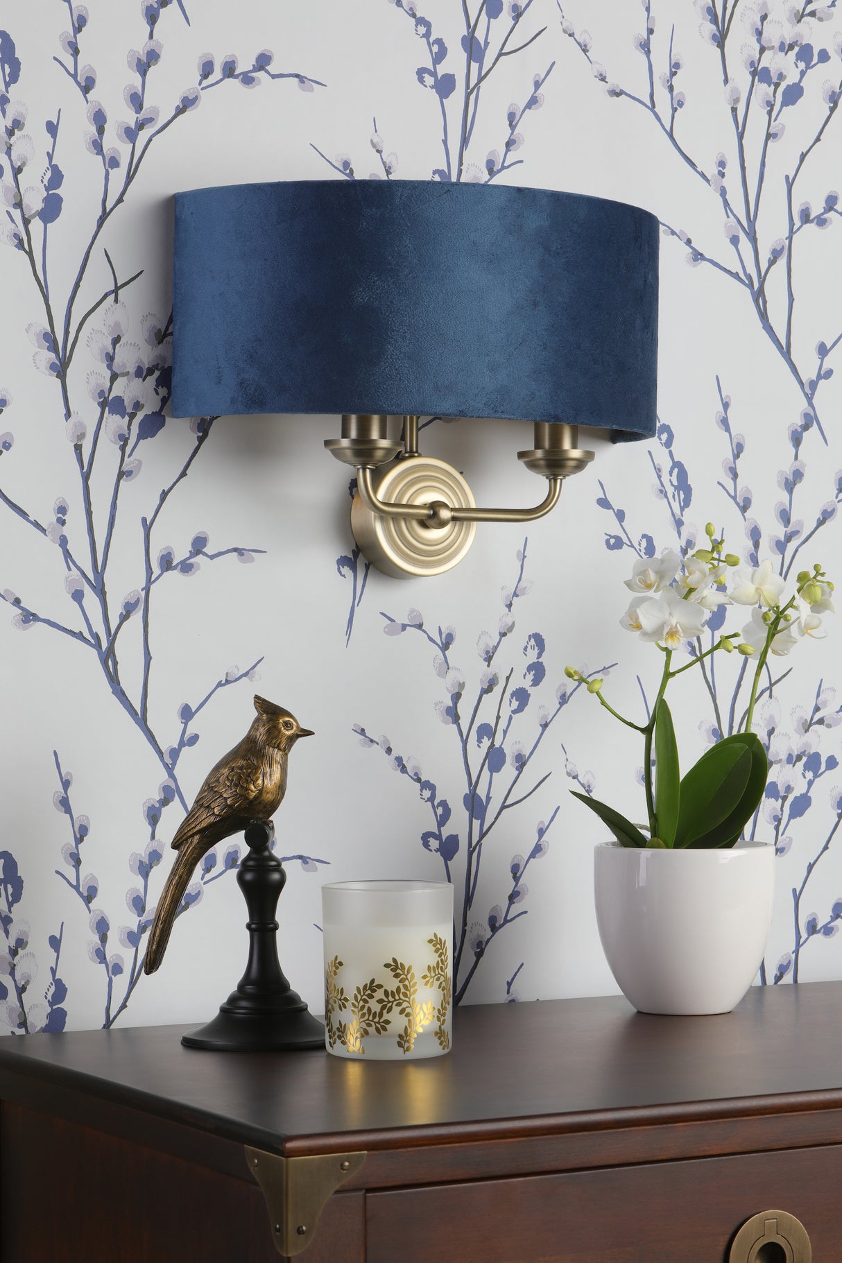 Decorative wooden cabinet with floral wallpaper, brass bird sculpture, frosted candle, white orchid, and blue wall sconce