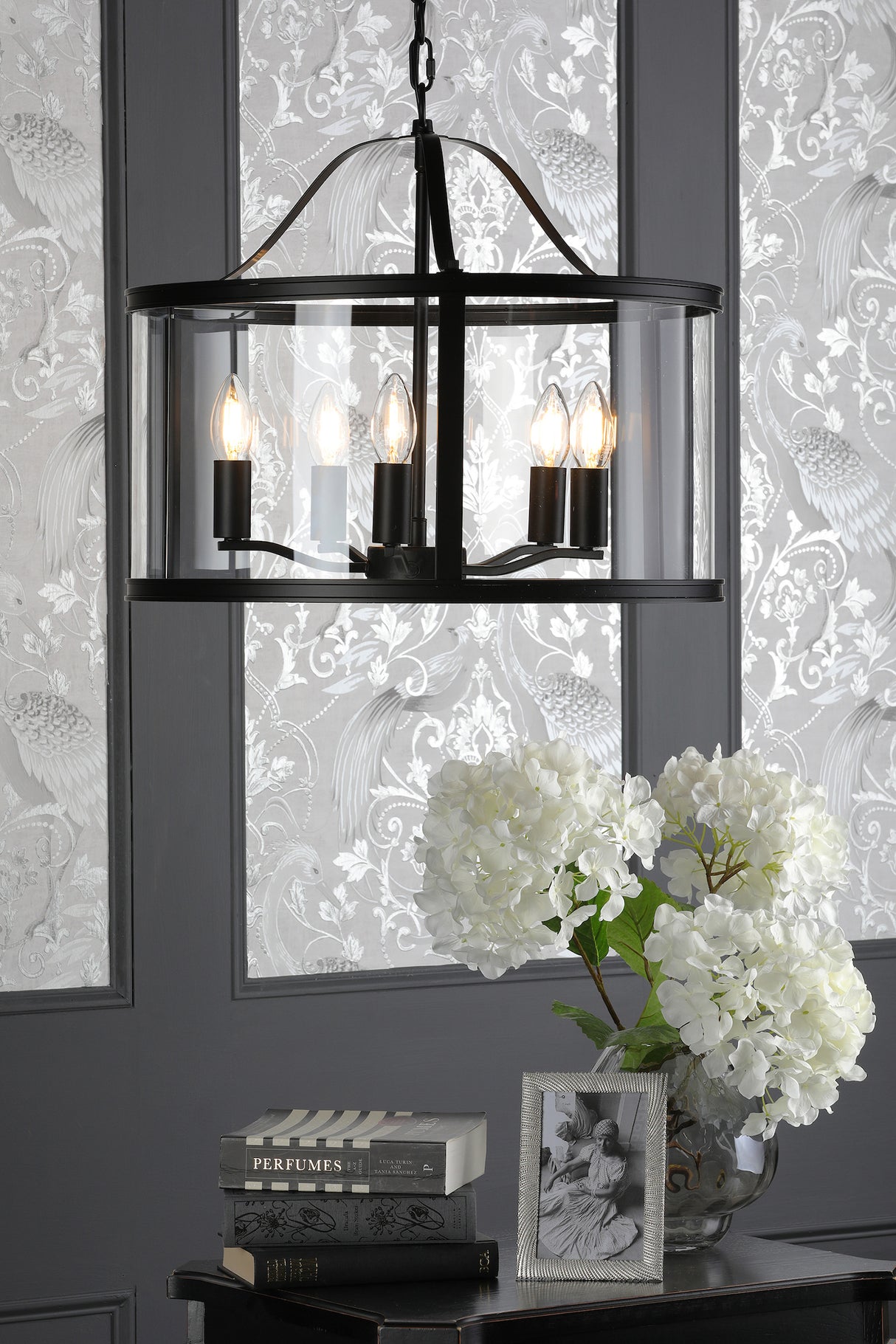 Black metal and glass chandelier with six lit bulbs over table with white hydrangeas, books, and framed photo