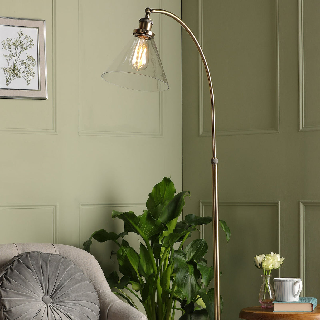 Modern brass floor lamp with clear glass shade beside green leafy plant and gray round pillow on beige chair