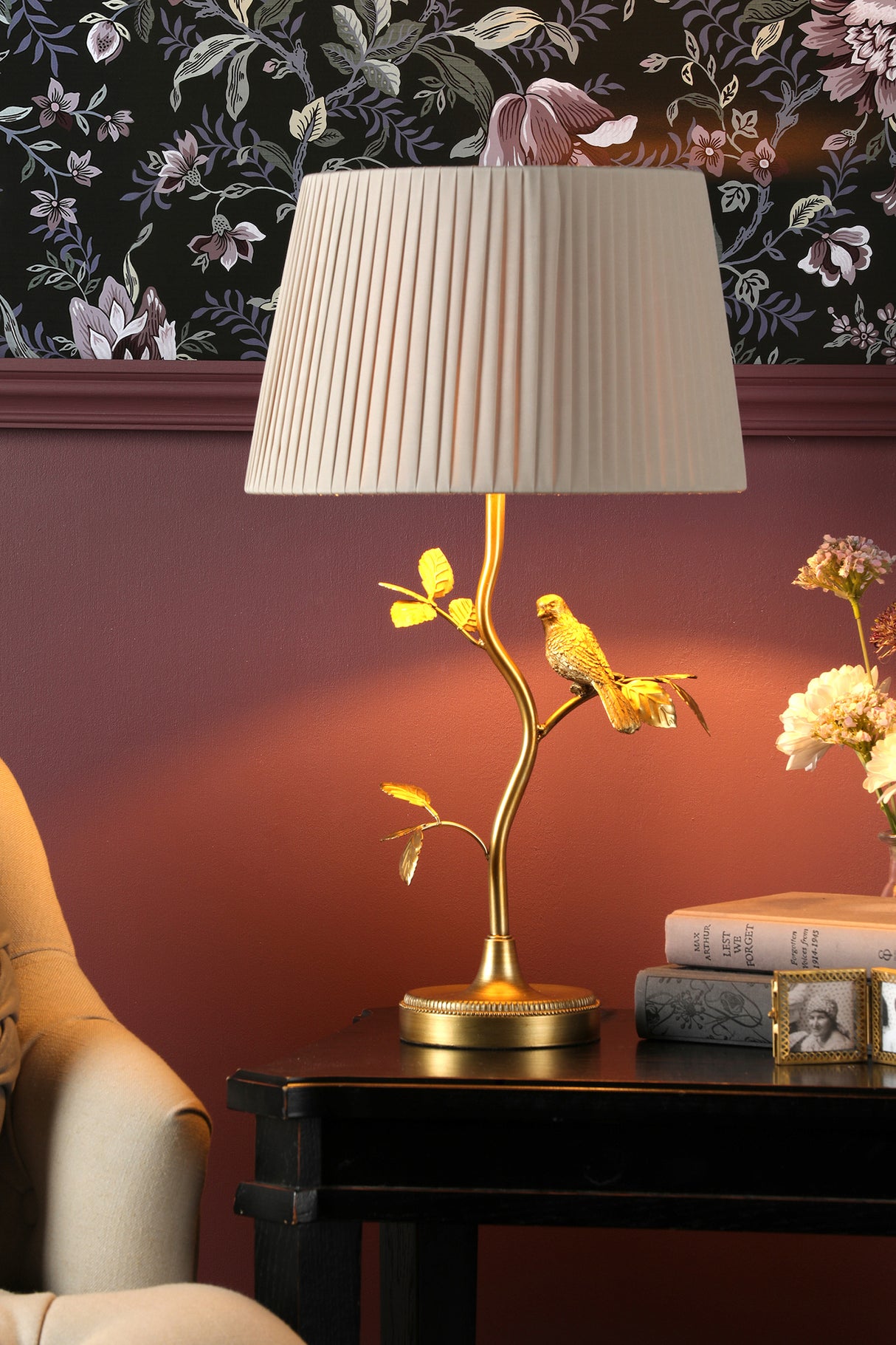 Elegant gold bird table lamp with pleated beige shade on black side table against floral wallpaper