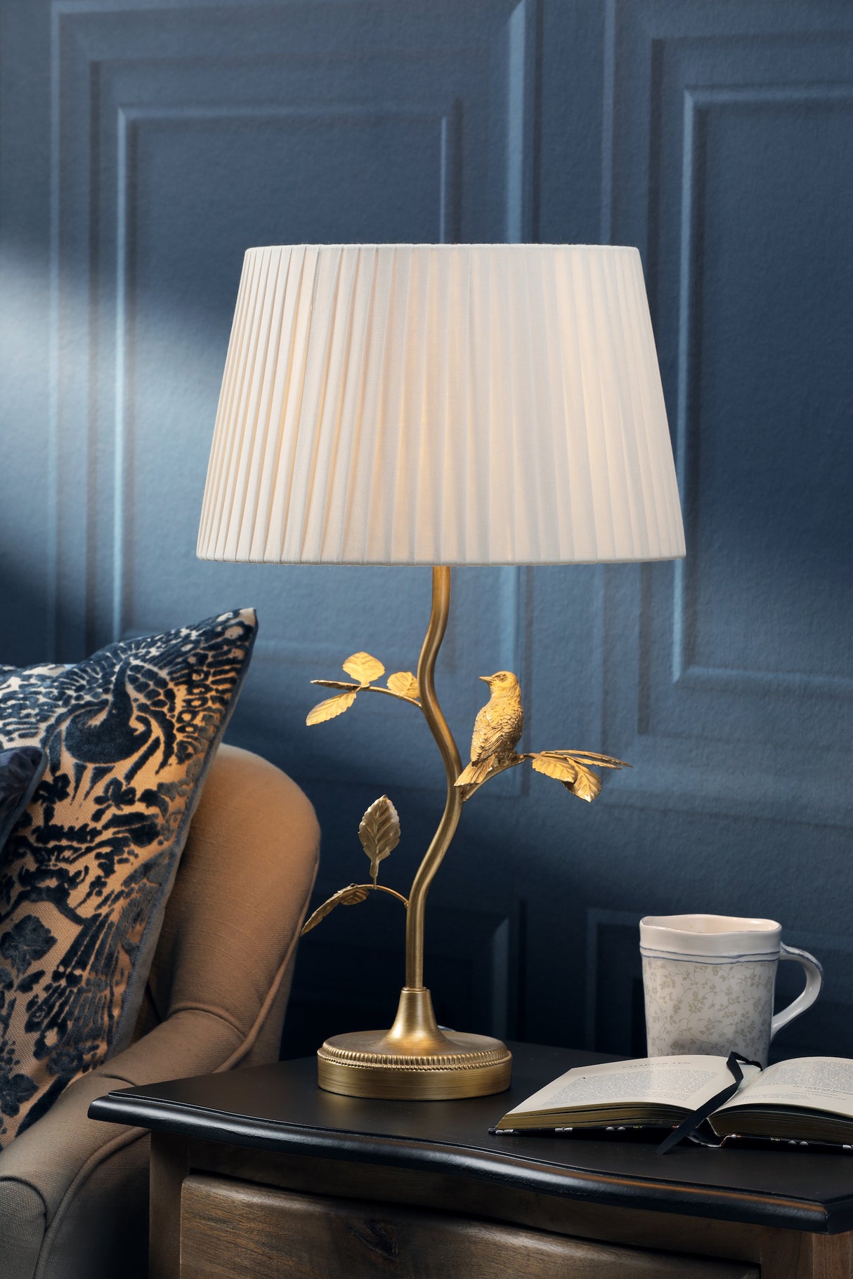 Elegant table lamp with pleated white shade and gold bird design on wooden side table with book and mug