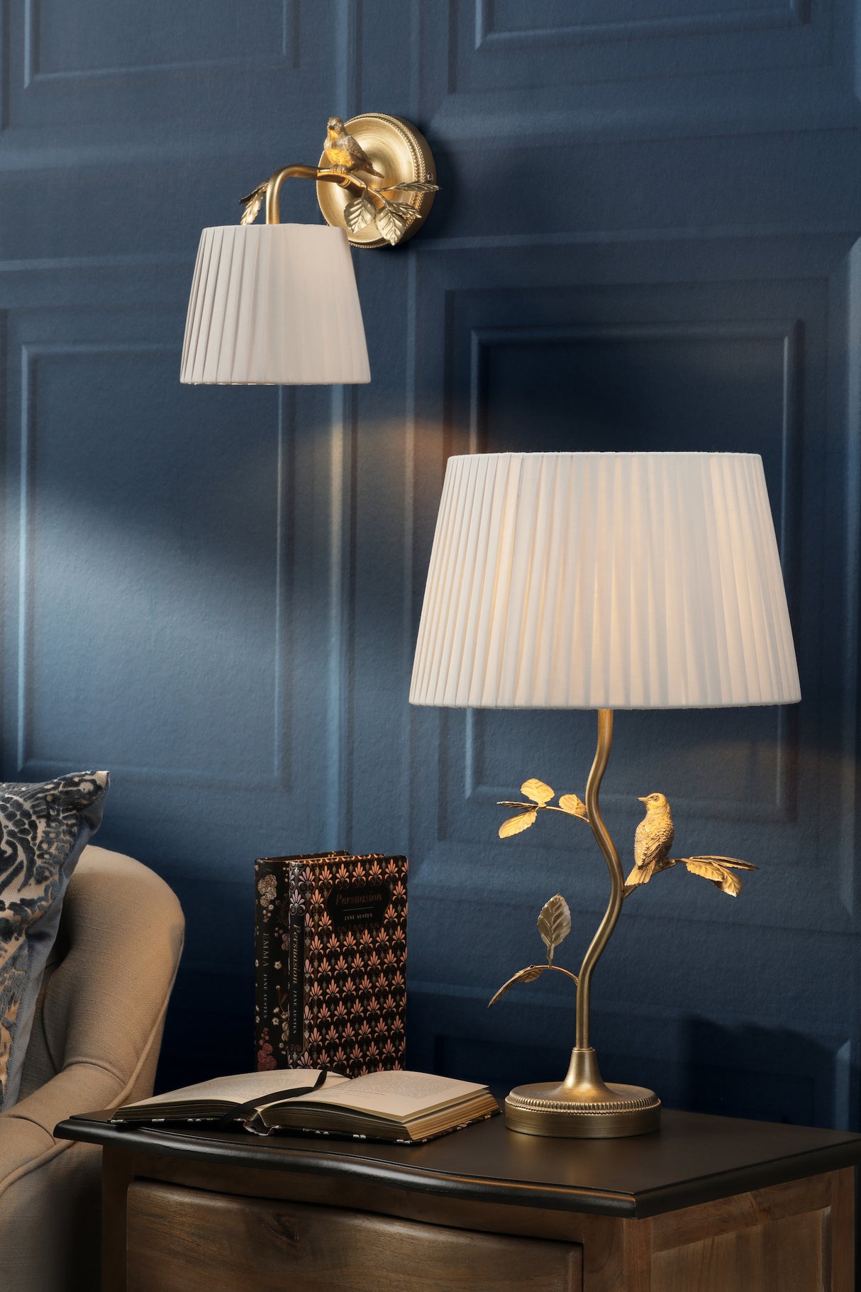 Elegant white lampshades on gold bird and leaf decor wall sconce and table lamp on dark blue paneled wall