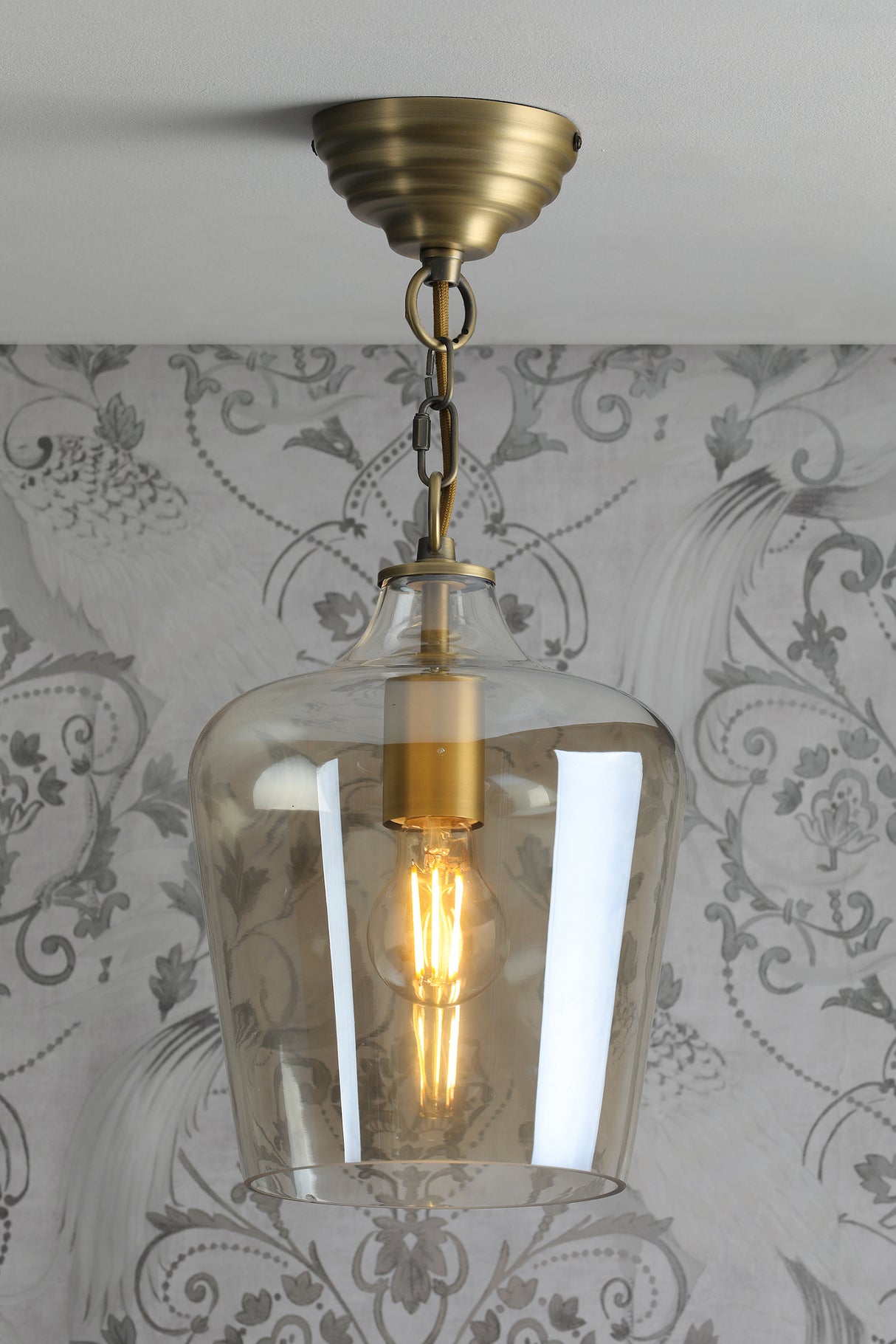 Brass ceiling pendant light with clear glass shade and vintage-style filament bulb against patterned wallpaper