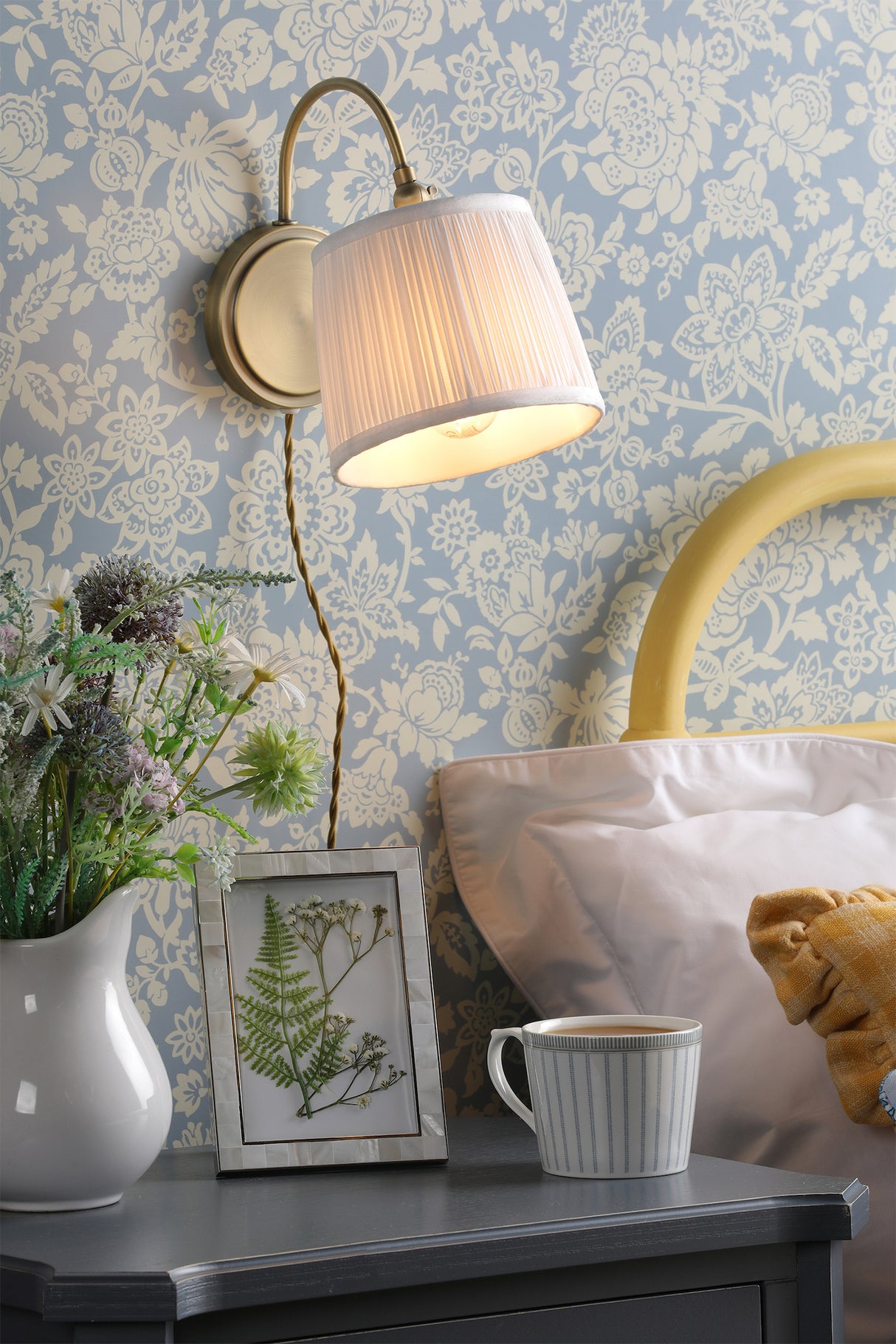 Cozy bedside table with floral wallpaper, wall lamp, vase of flowers, framed pressed plants, and cup of tea
