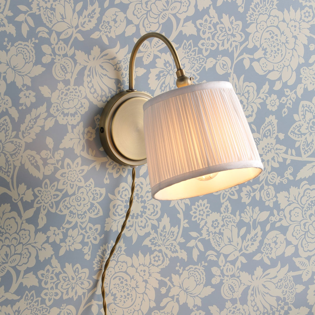 Brass wall sconce with pleated white lampshade mounted on blue and cream floral wallpaper