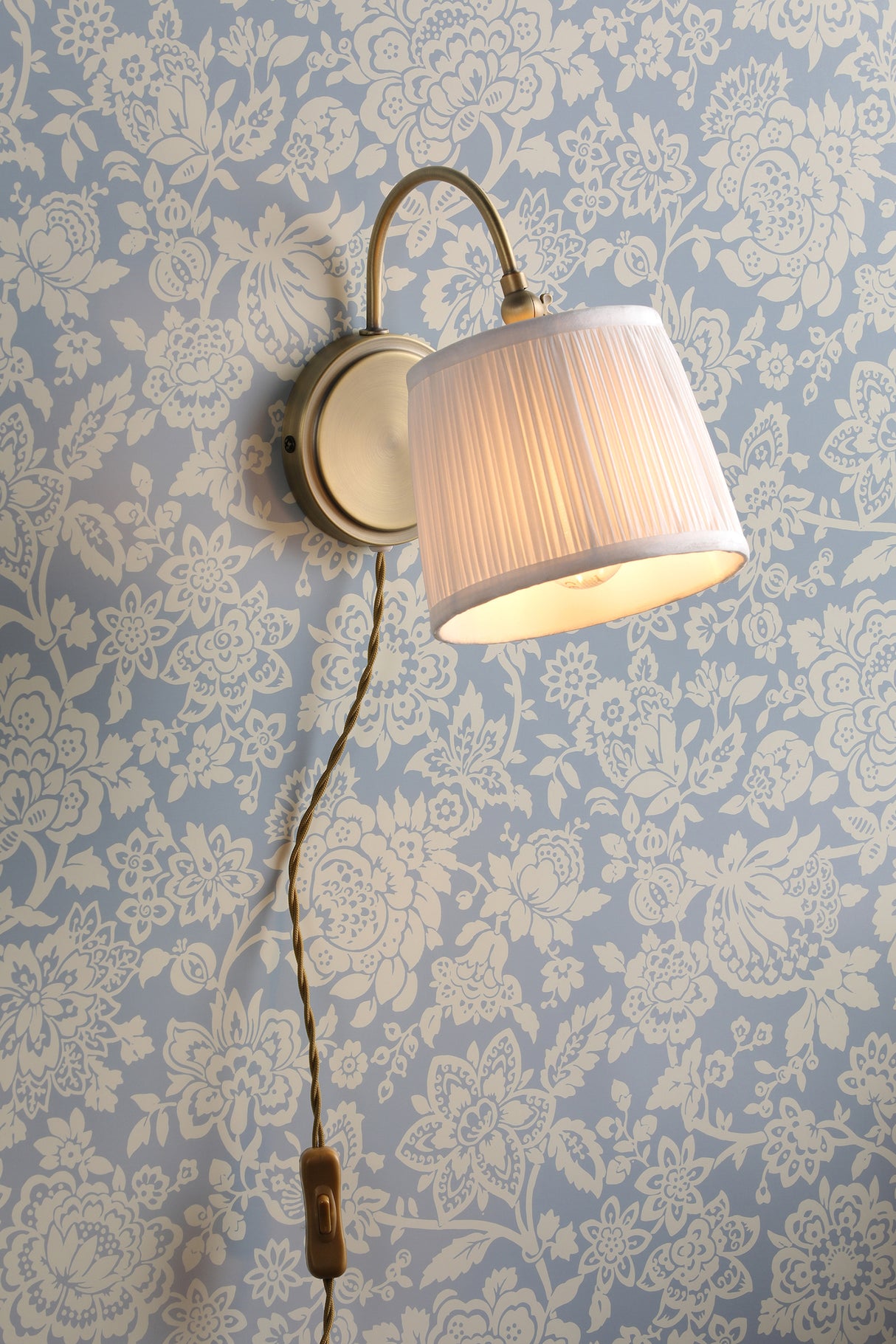 Brushed brass wall sconce with pleated cream lampshade on blue floral wallpaper, vintage-style cord switch