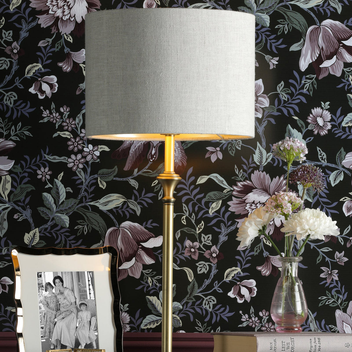 Gold floor lamp with gray shade beside photo frame and flowers in glass vase against floral wallpaper