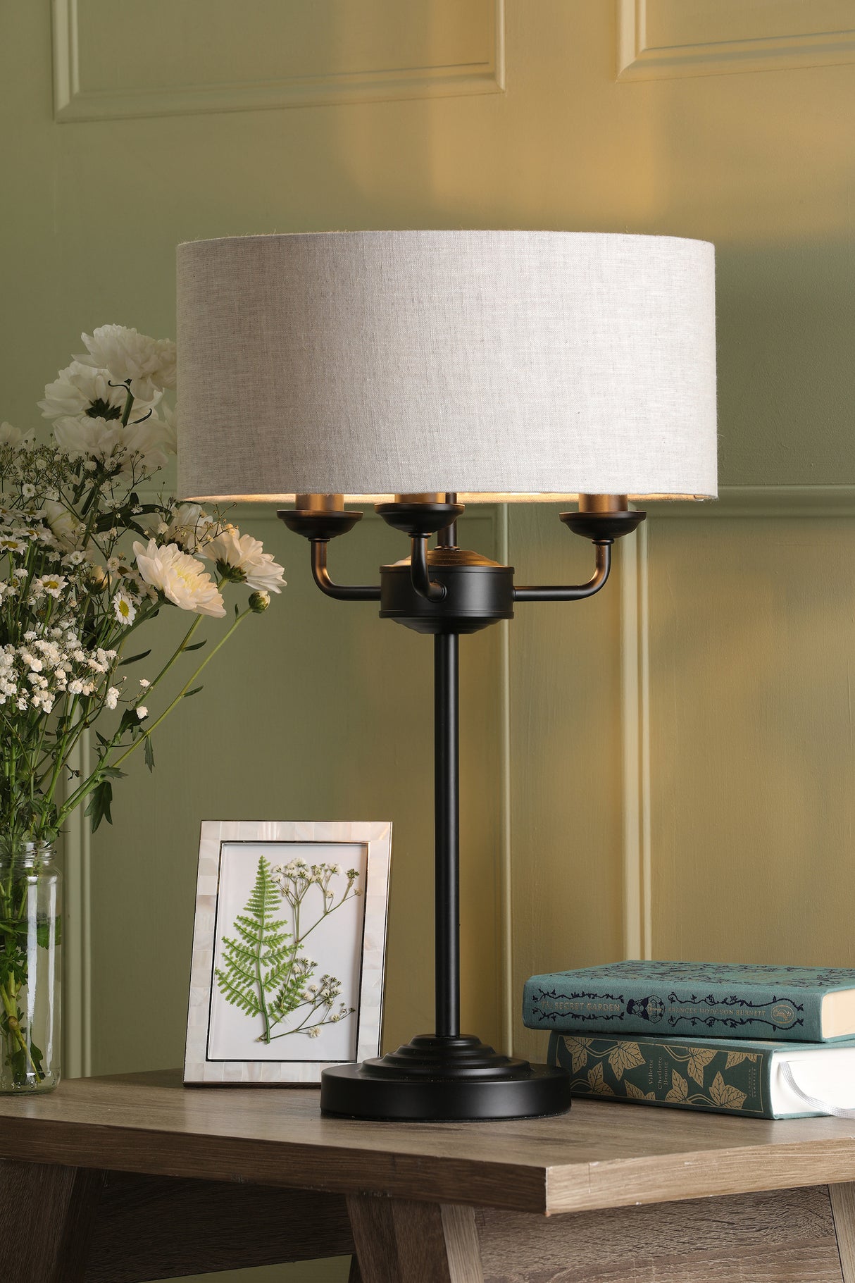 Black metal table lamp with cream fabric shade on wooden desk with flowers, framed botanical art, and stacked books
