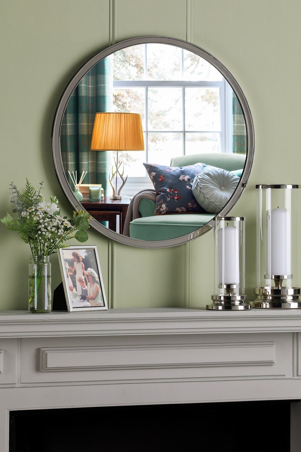 Round wall mirror reflecting green armchair with floral and round pillows beside window with plaid curtains