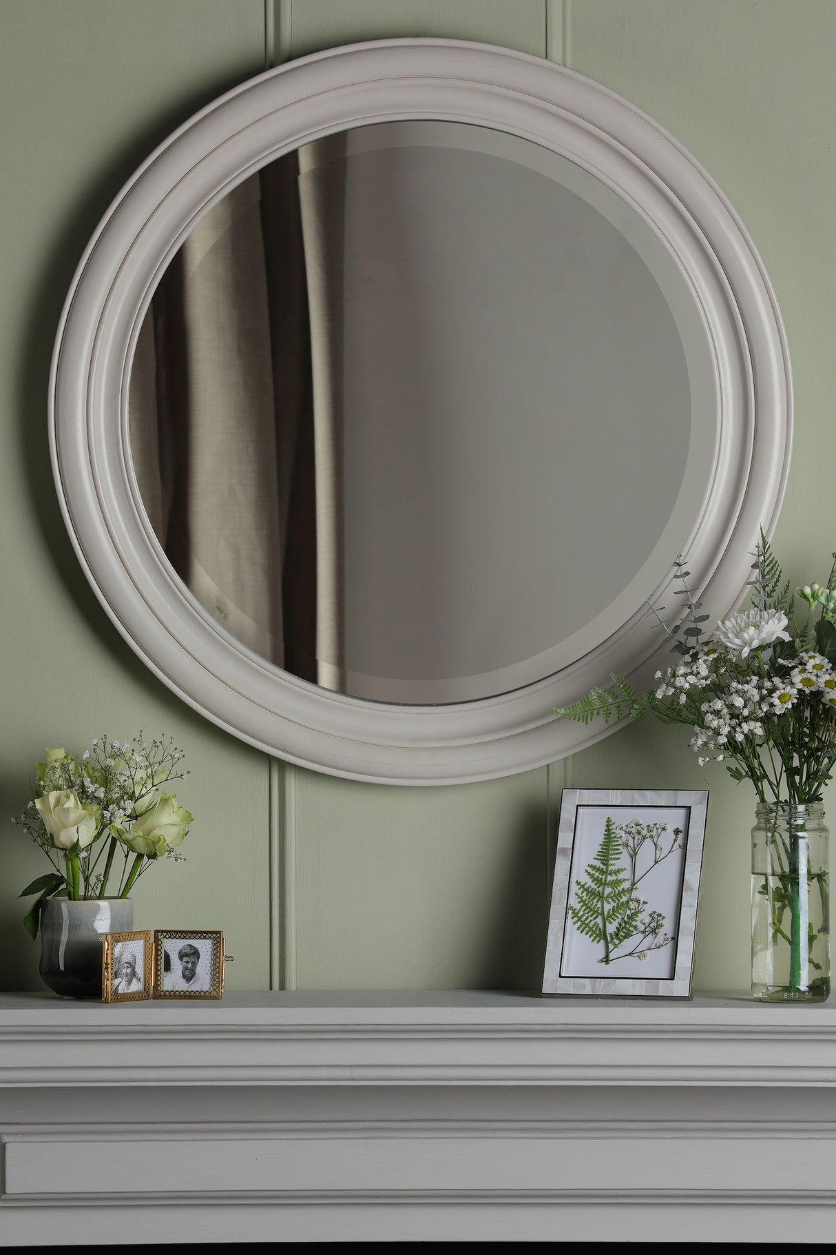 Round white framed mirror above light gray mantel with flowers, photo frame, and fern art