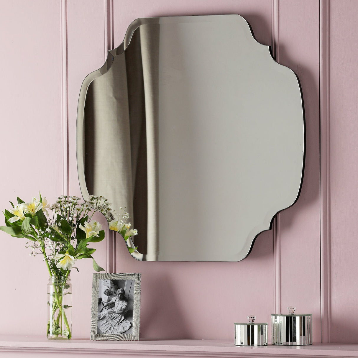 Decorative wall mirror with curved edges above pink mantel with flowers, photo frame, and silver canisters