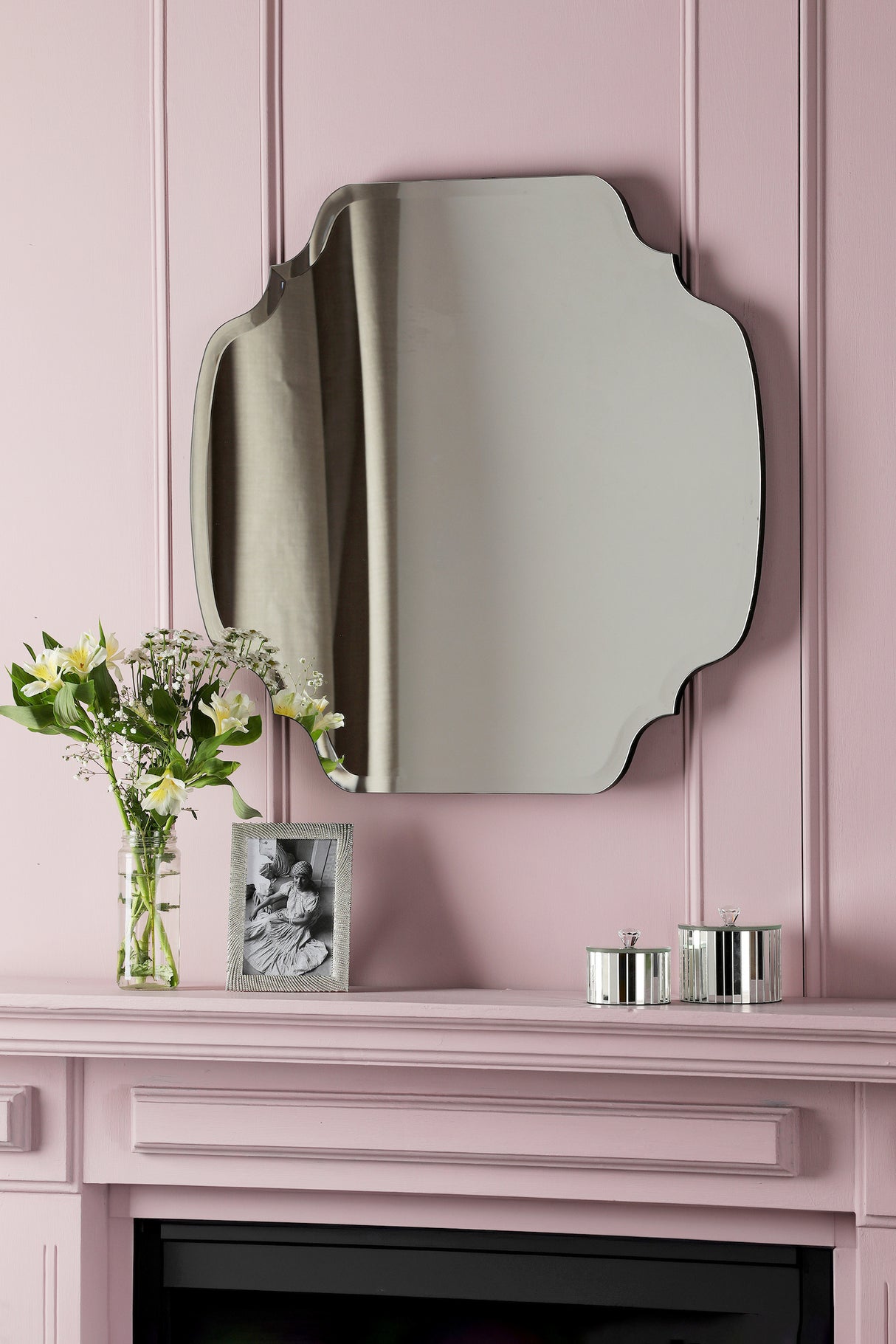 Decorative vintage mirror on pink wall above mantel with flowers, photo frame, and silver containers