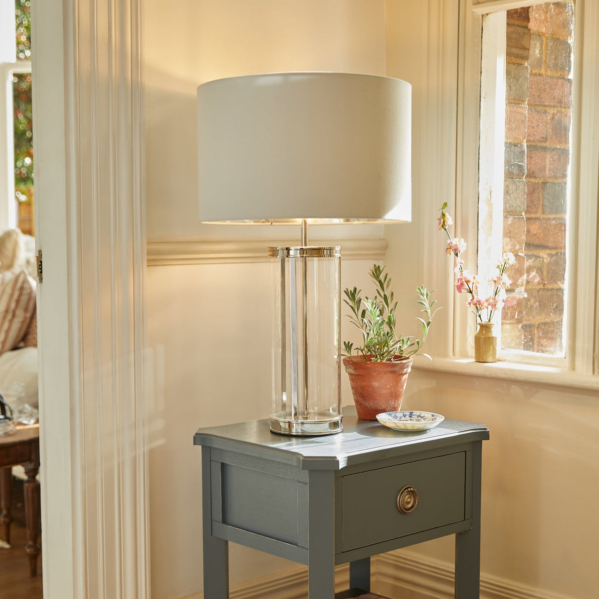 A Laura Ashley Harrington Large Table Lamp with a polished nickel finish, placed on a side table, with a white lampshade, in a room with a window and a plant.