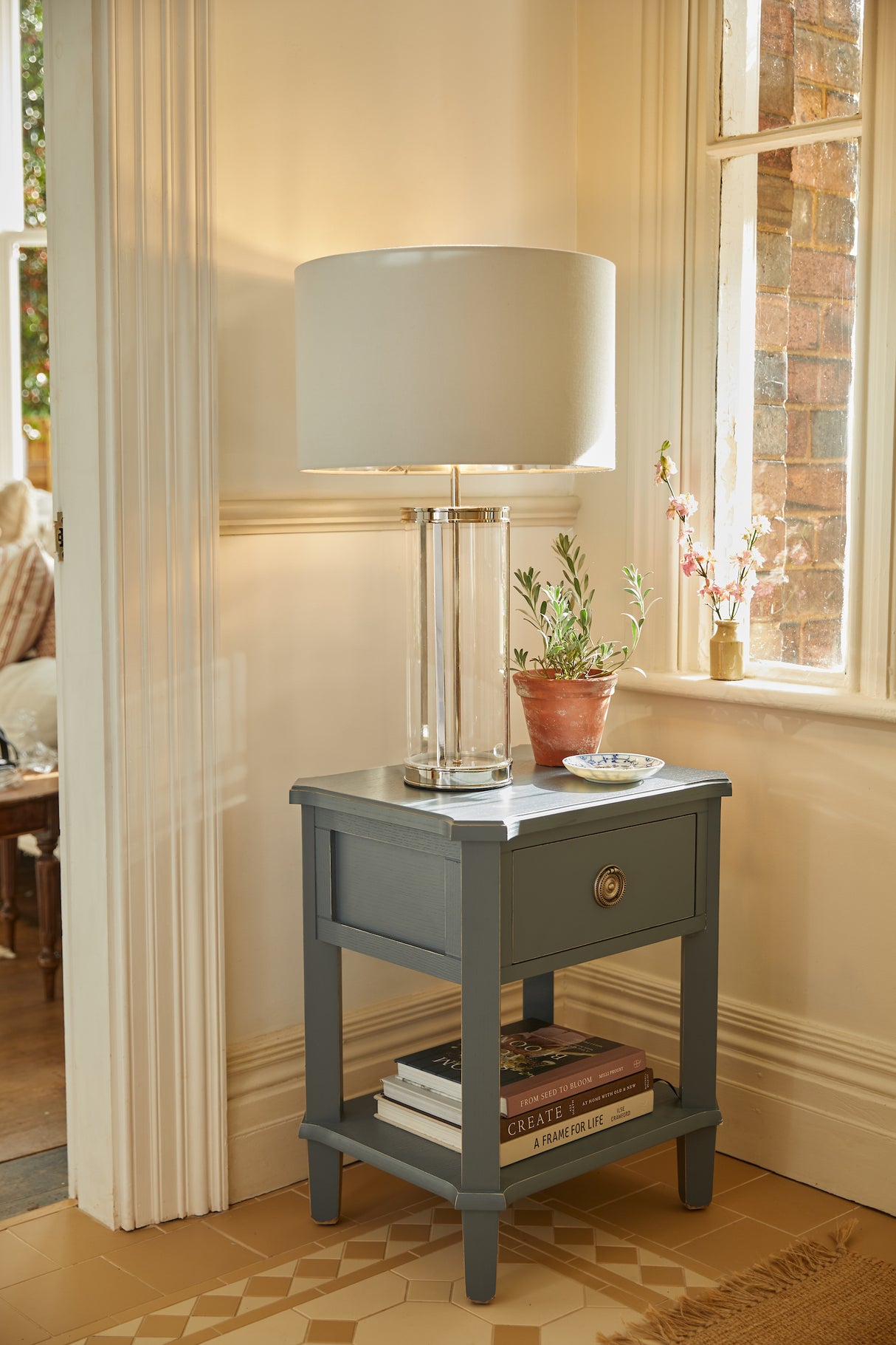 Gray wooden side table with glass lamp, potted plant, books, and floral vase by window in cozy room