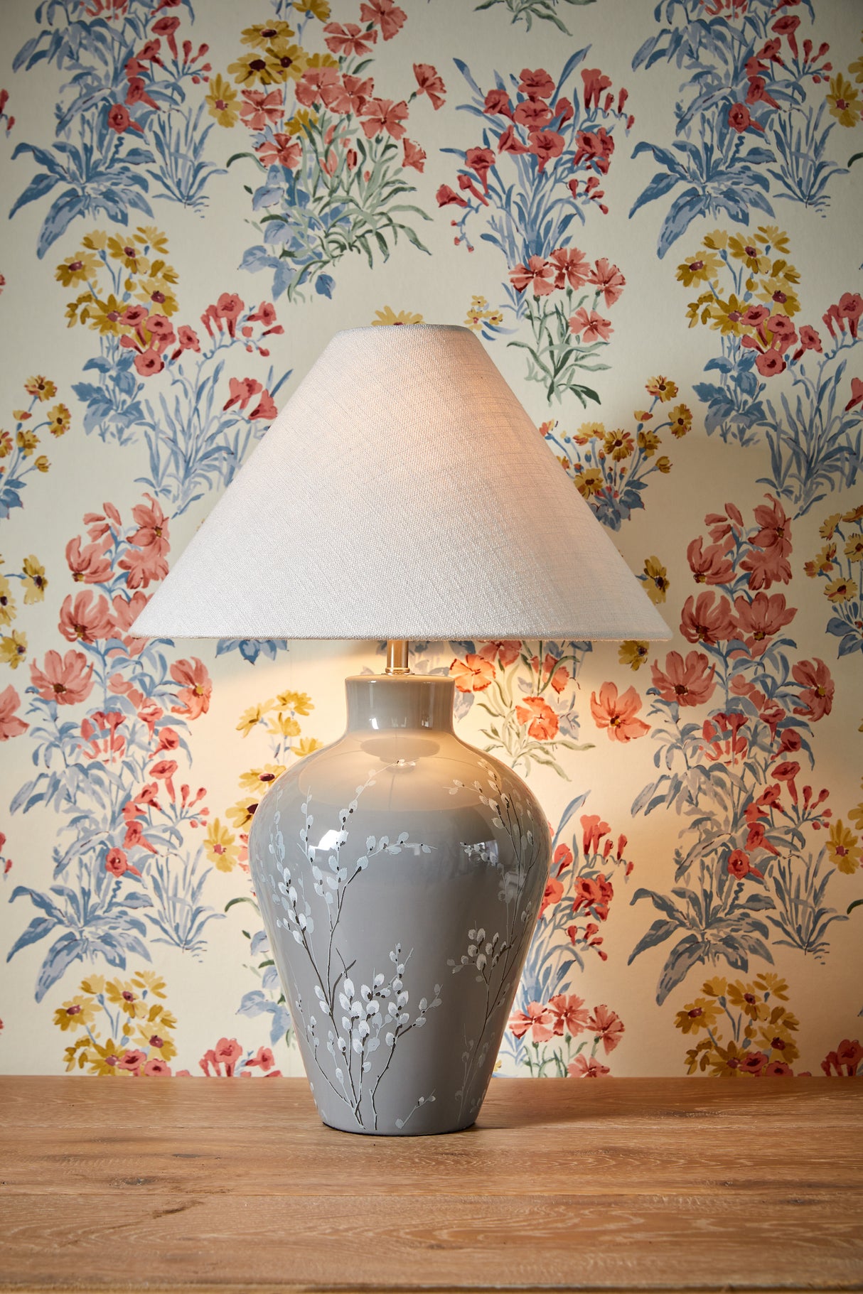 Ceramic table lamp with white floral patterns and beige lampshade on wood table, floral wallpaper background