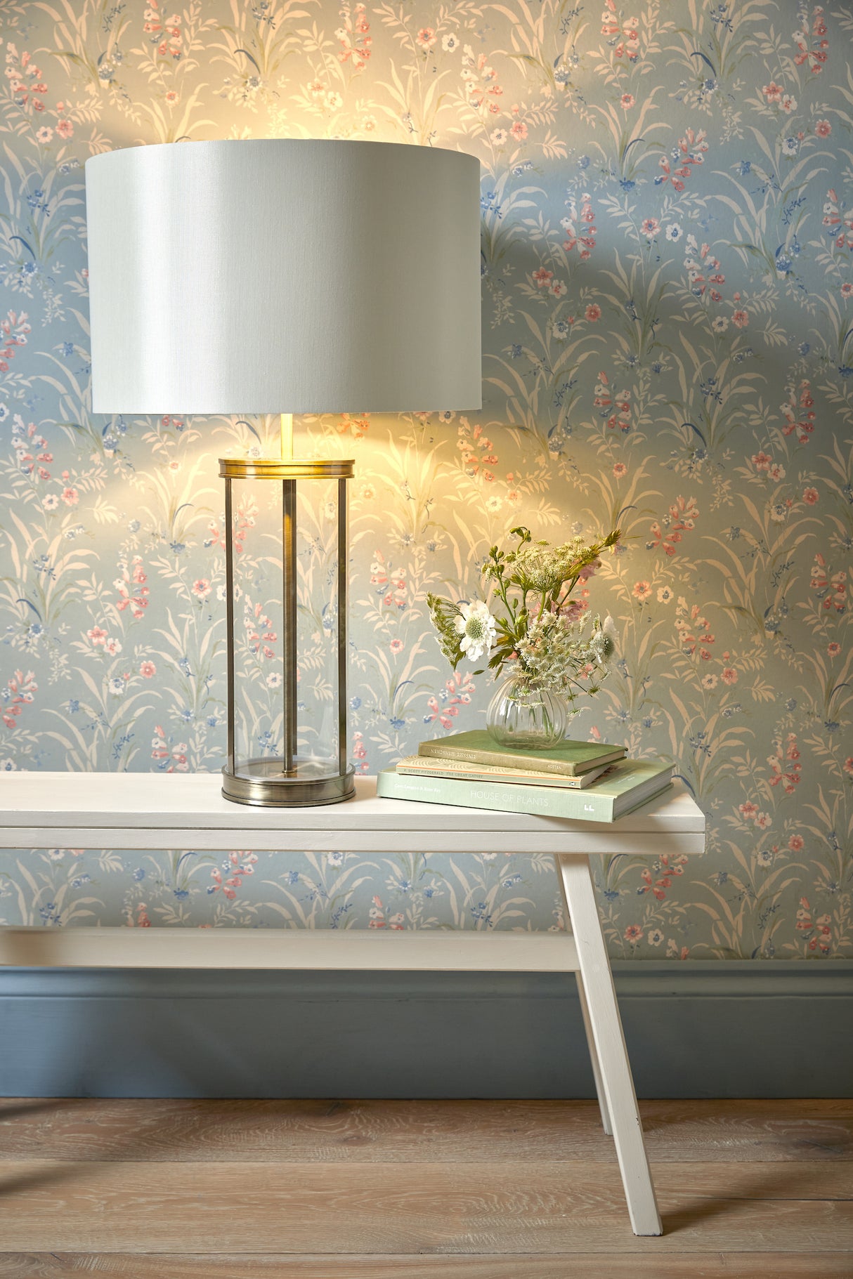 Modern table lamp with glass base and white shade on white bench with books and small flower vase