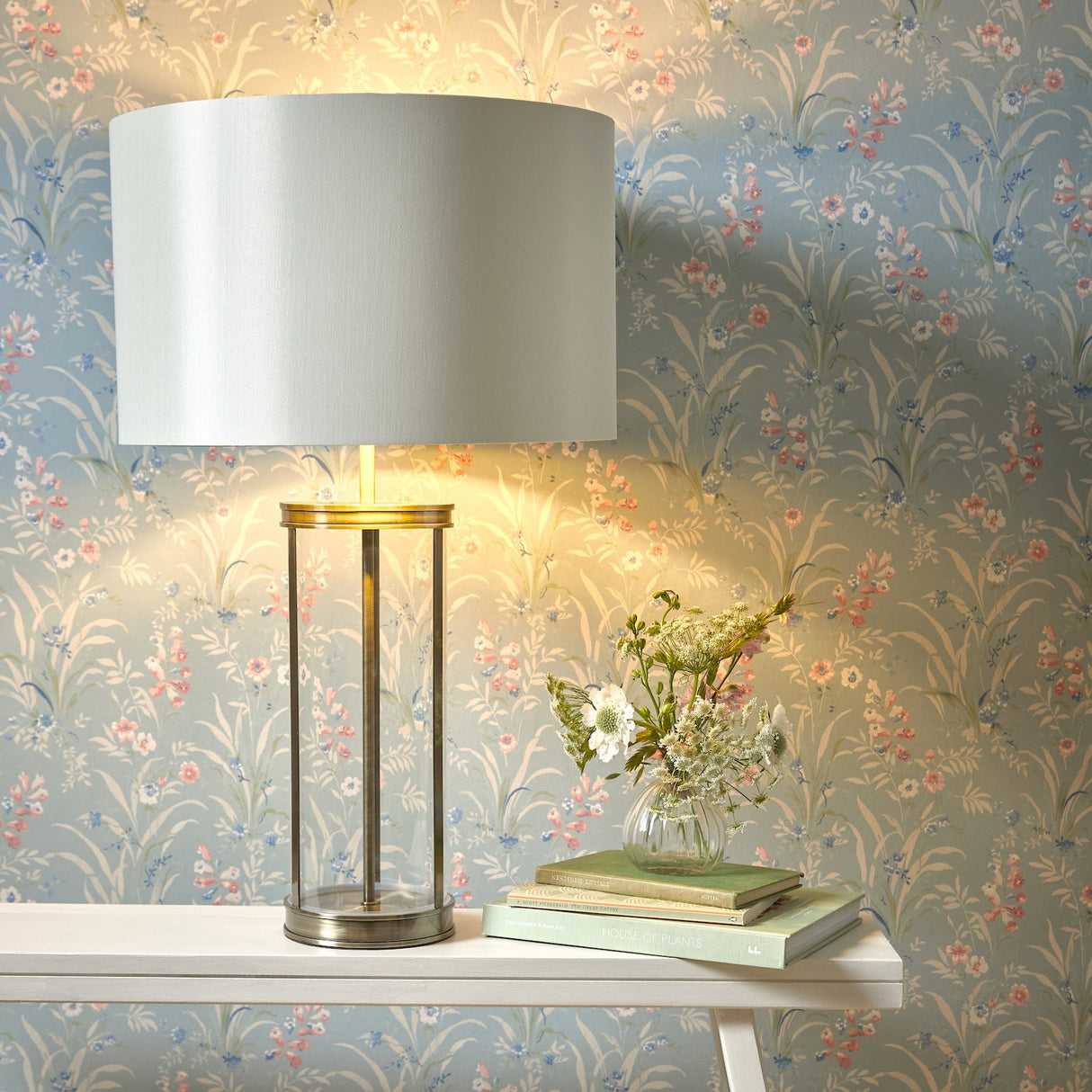 A Laura Ashley Harrington Large Table Lamp with antique brass finish and a white lampshade, displayed against a patterned wallpaper background.
