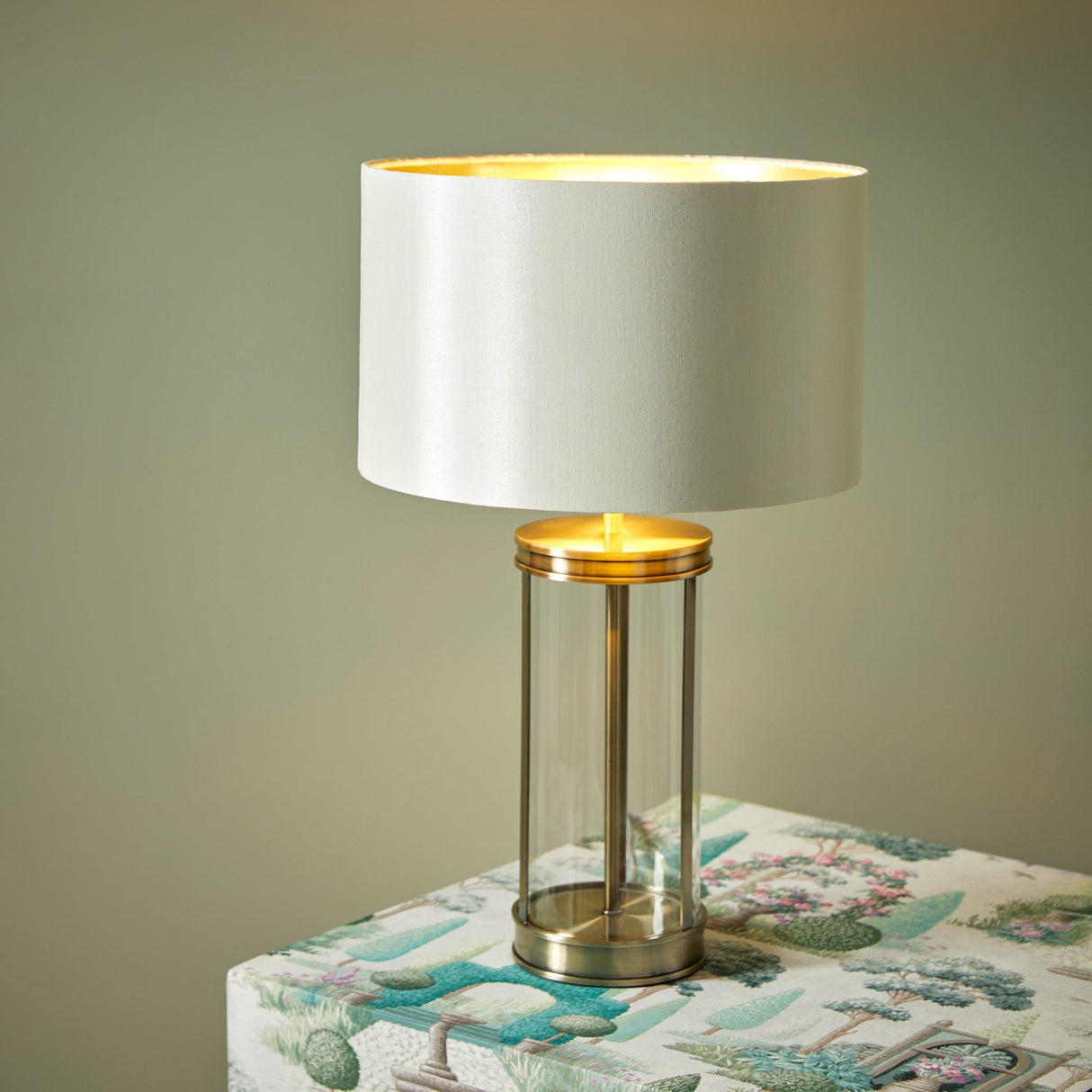 A Laura Ashley Harrington Small Table Lamp with antique brass finish, a clear cylindrical base, and a white lampshade, placed on a table with a patterned tablecloth.