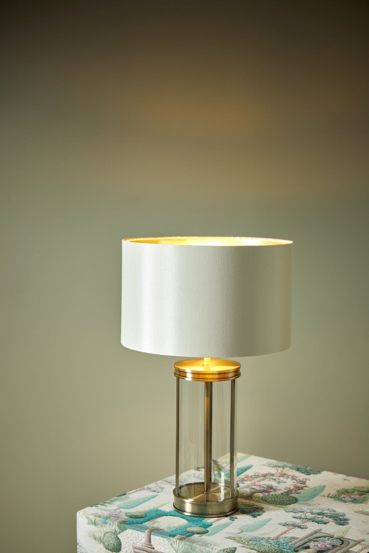 Modern table lamp with a white fabric shade and transparent cylindrical glass base on floral-patterned surface