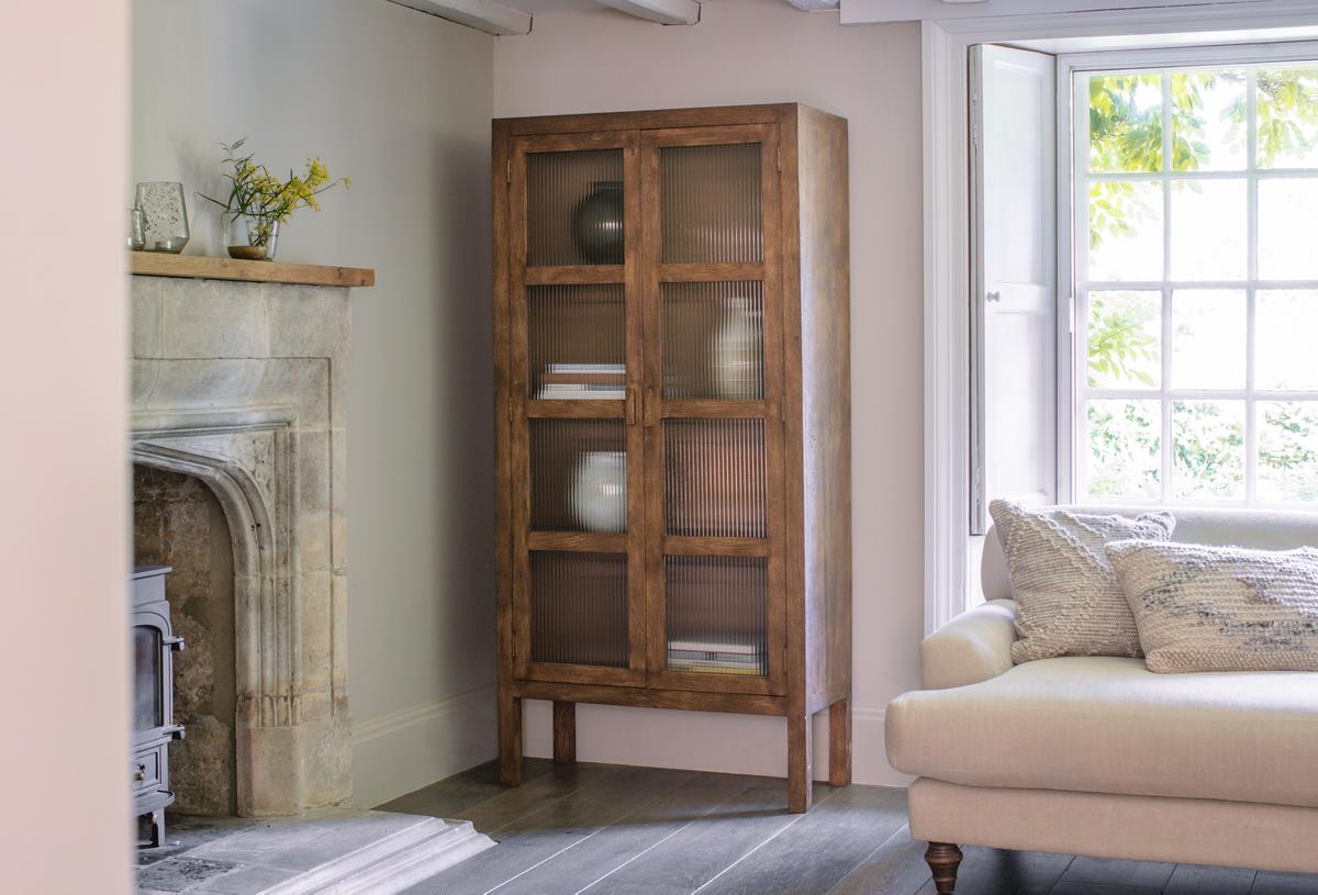 A walnut brown Nkuku Mendani Mango Wood & Fluted Glass Cabinet with two doors, placed in a room near a white sofa and a traditional fireplace.