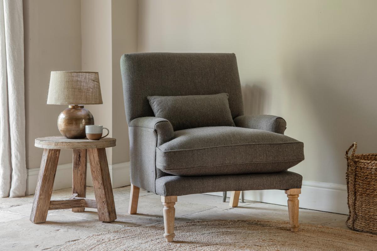 A charcoal gray Nkuku Nalla Wool Armchair with a subtle self herringbone pattern, placed in a room with a wooden side table, a lamp, and a basket.