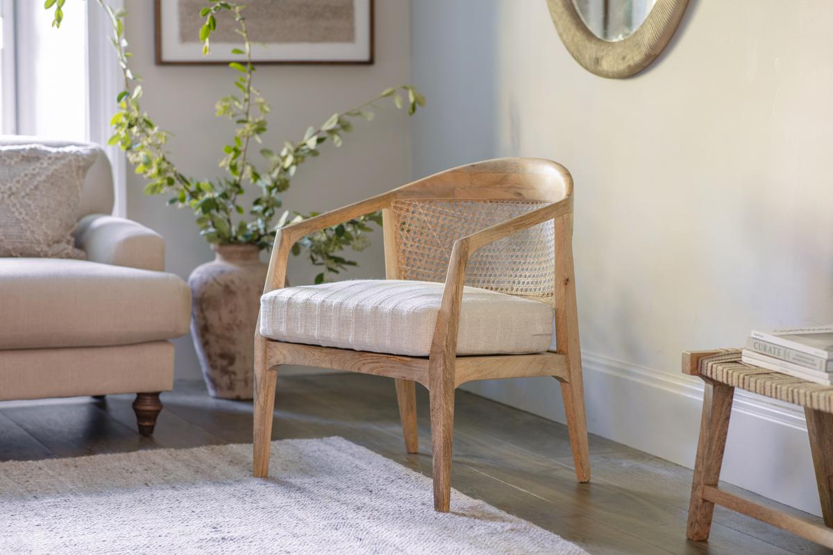 A natural-colored Nkuku Nabhi Mango Wood & Cane Occasional Chair with a white cushion, placed in a room setting with a plant and a mirror in the background.