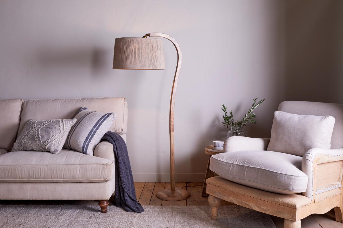 A tall, sleek Nkuku Namira Mango Wood Floor Lamp with a natural finish, pictured in a room setting with a woven lampshade not included.