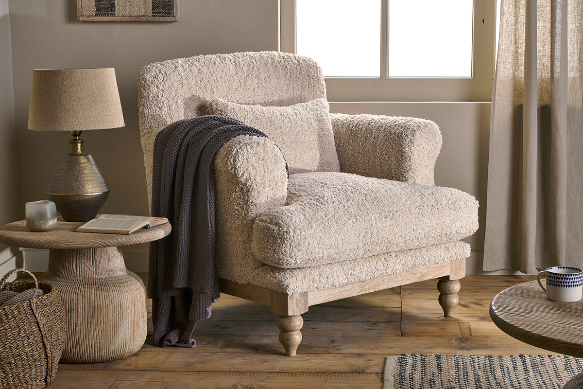A beige Nkuku Otas Teddy Cotton Armchair with a wooden frame, situated in a room with a lamp, curtains, and a side table.