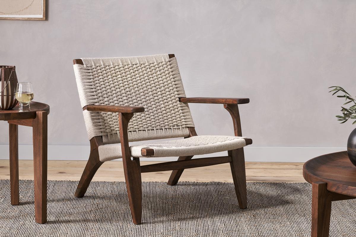 A beige Nkuku Surat Woven Lounger chair with Acacia Wood frame and hand-woven cotton rope, set in a room with a side table and plant.