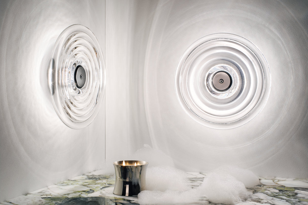 Modern circular glass wall lights creating ripple effects on white walls with silver candle and foam on marble surface