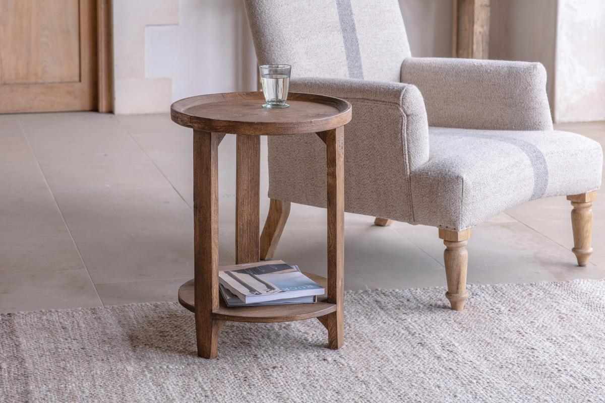 A walnut-finished Nkuku Triveni Mango Wood Side Table placed on a rug, with a glass of water on top and a chair in the background.