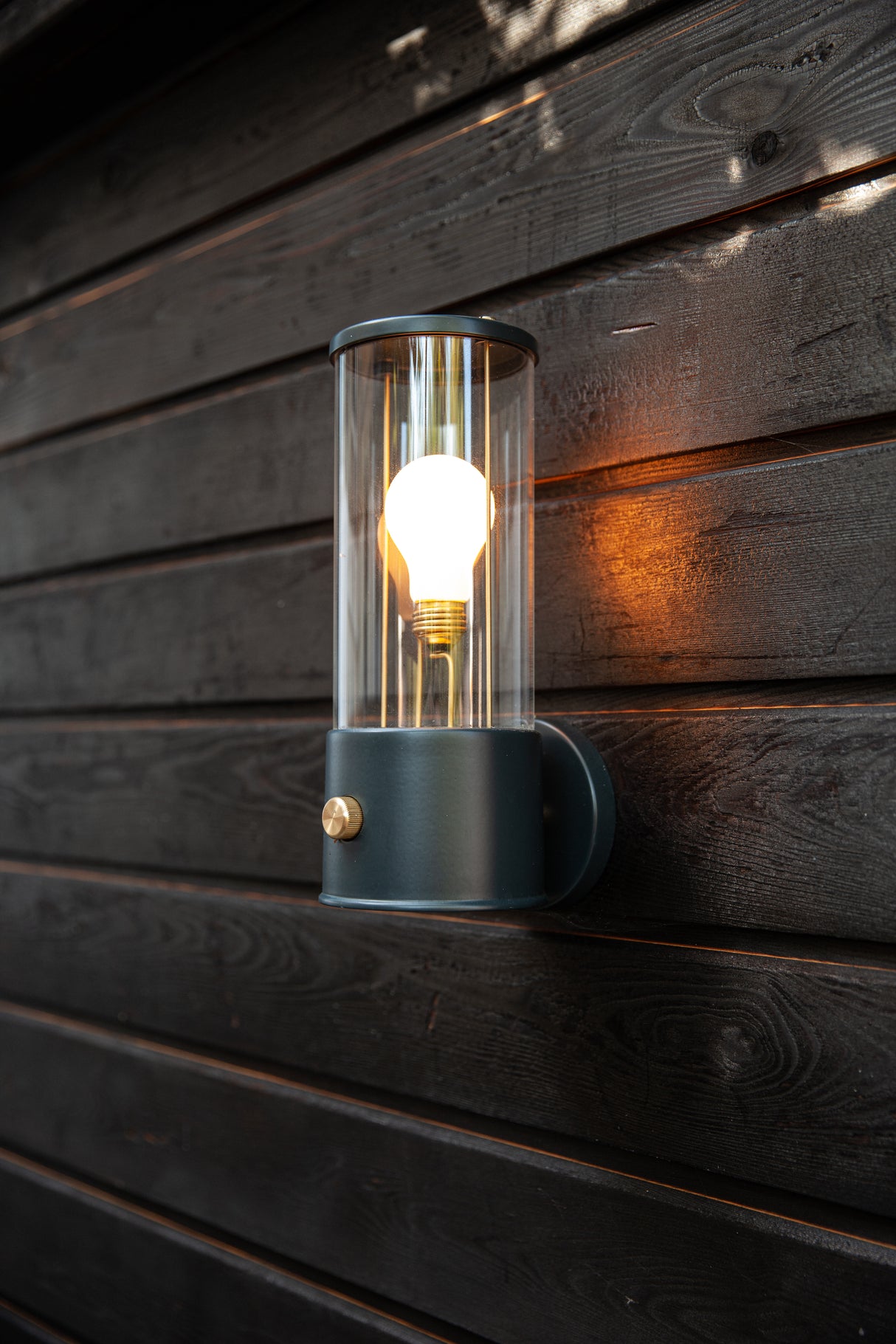 Modern black metal and glass wall light with glowing bulb mounted on dark wooden exterior wall