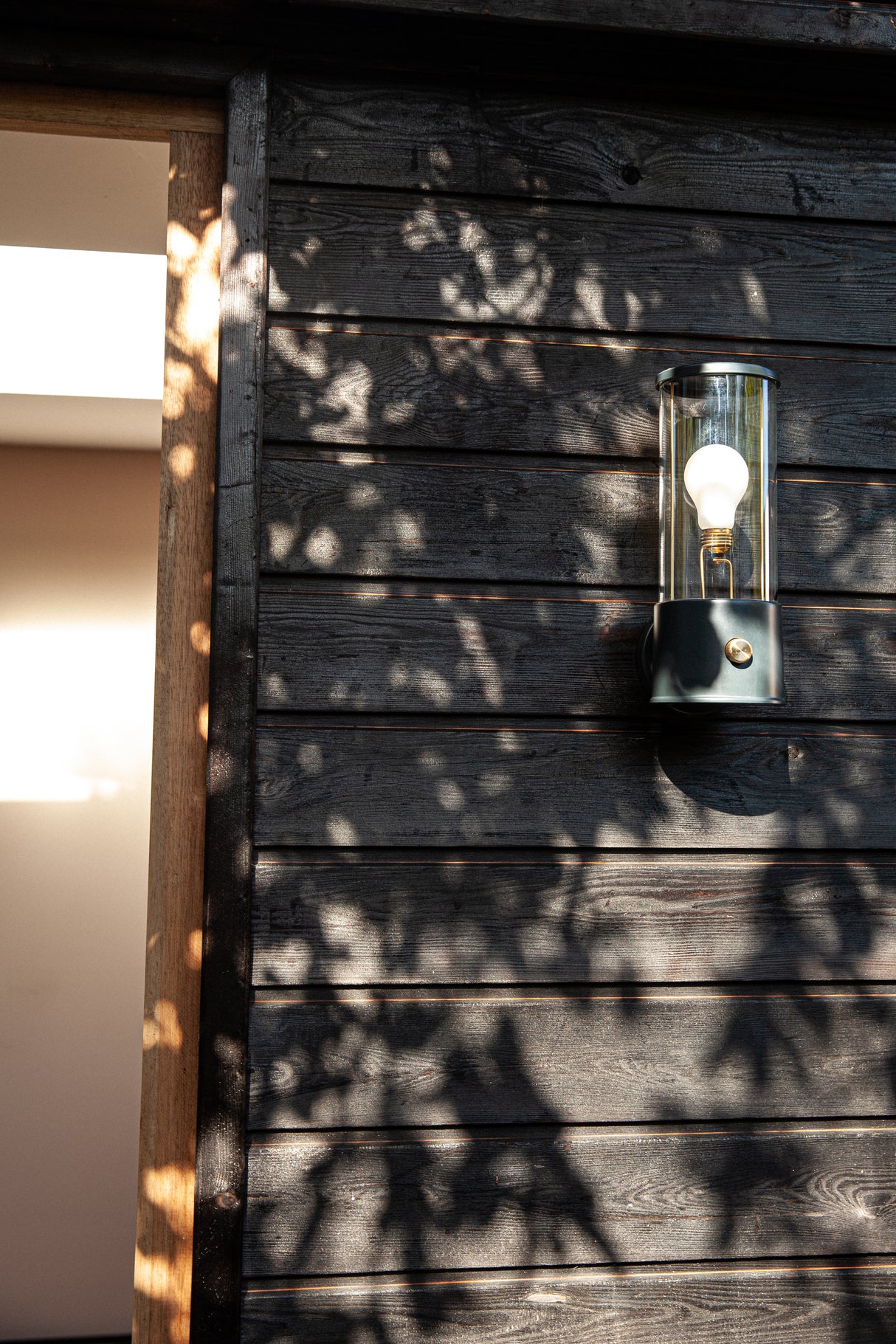 Modern Tala Muse wall light mounted on dark wooden exterior wall with dappled sunlight shadows